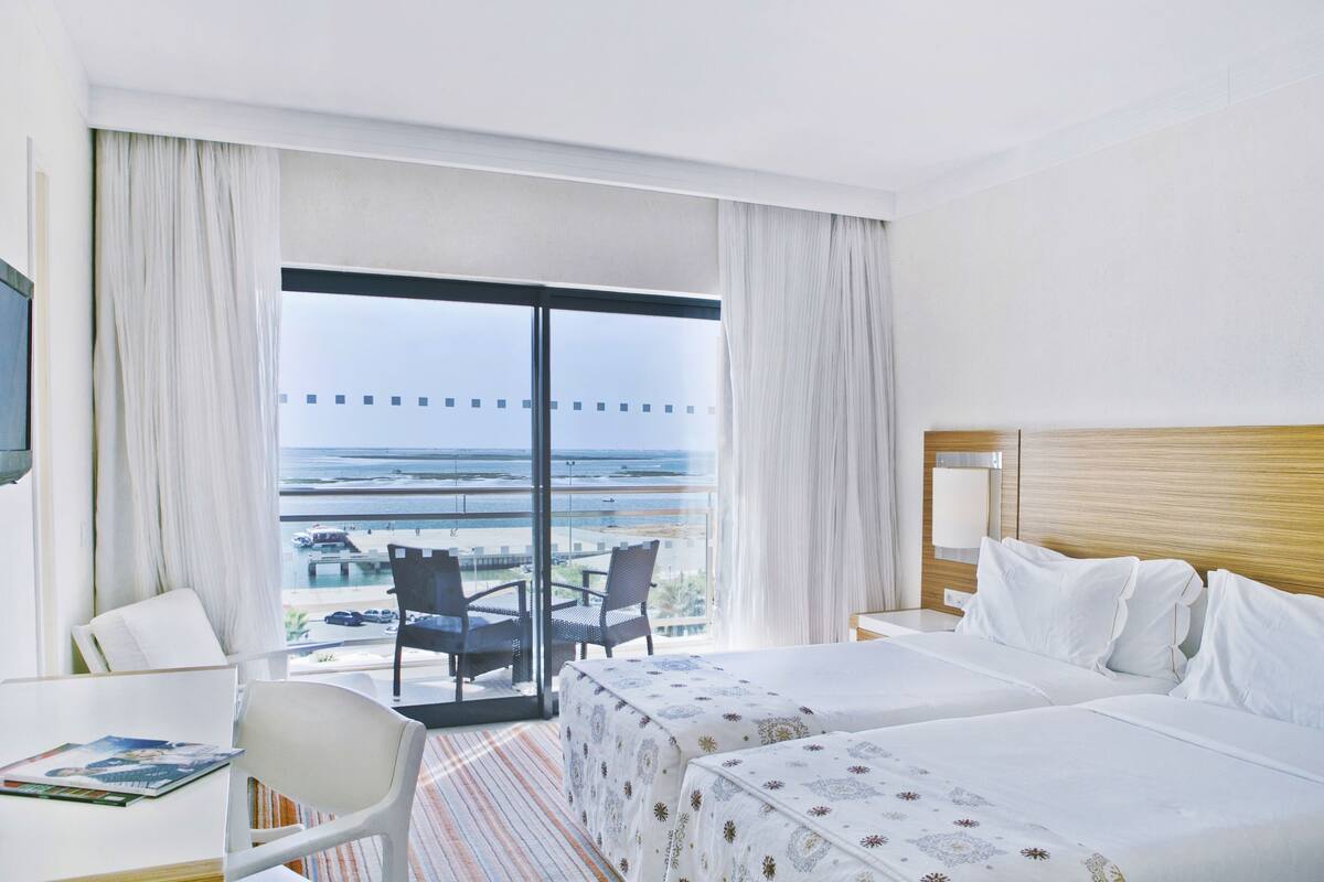 A hotel room with two beds, a desk, a chair, and a balcony with a view of the ocean.