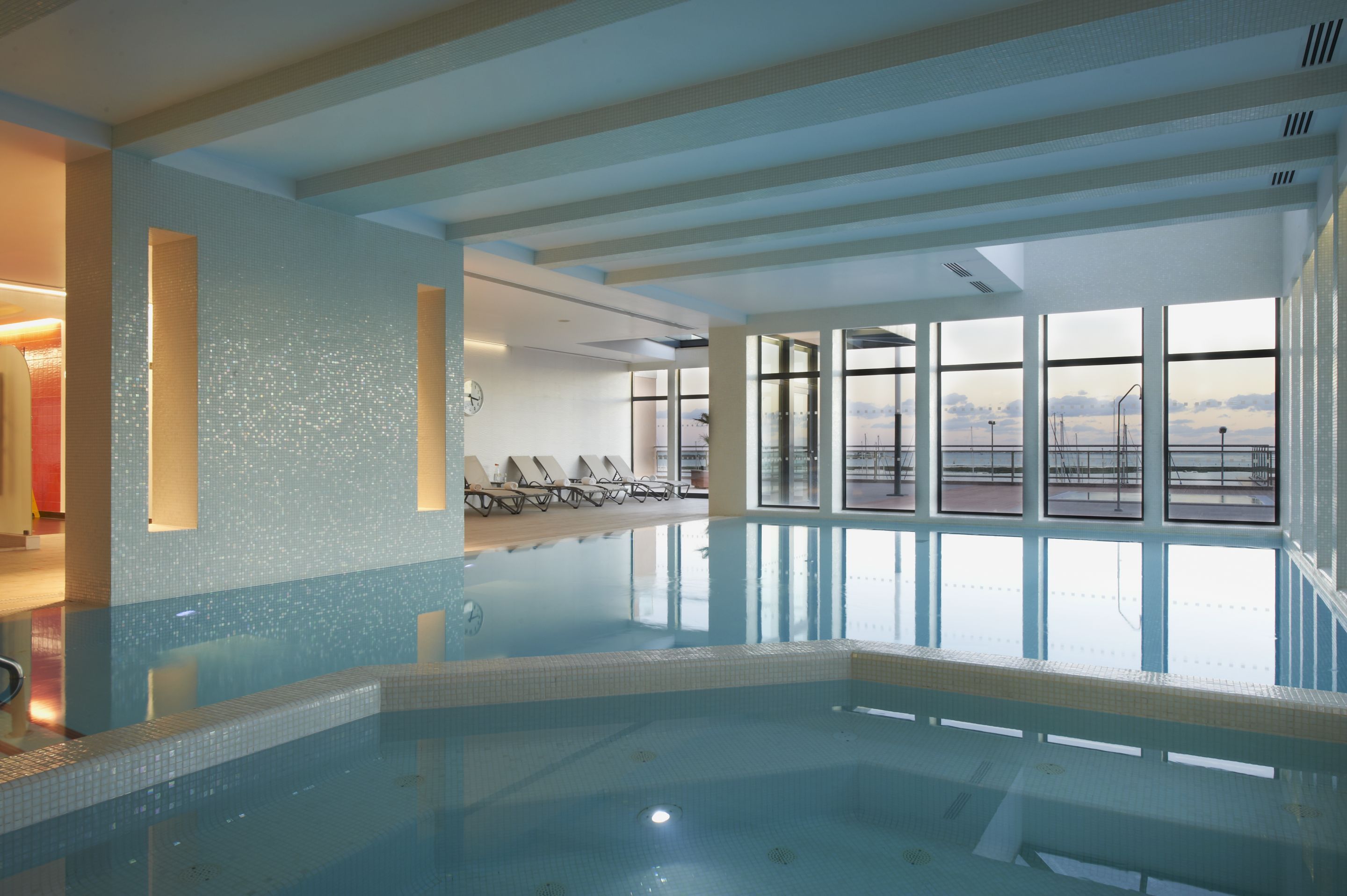 An indoor swimming pool with clear blue water, a tiled floor, and a view of the sky through large windows.