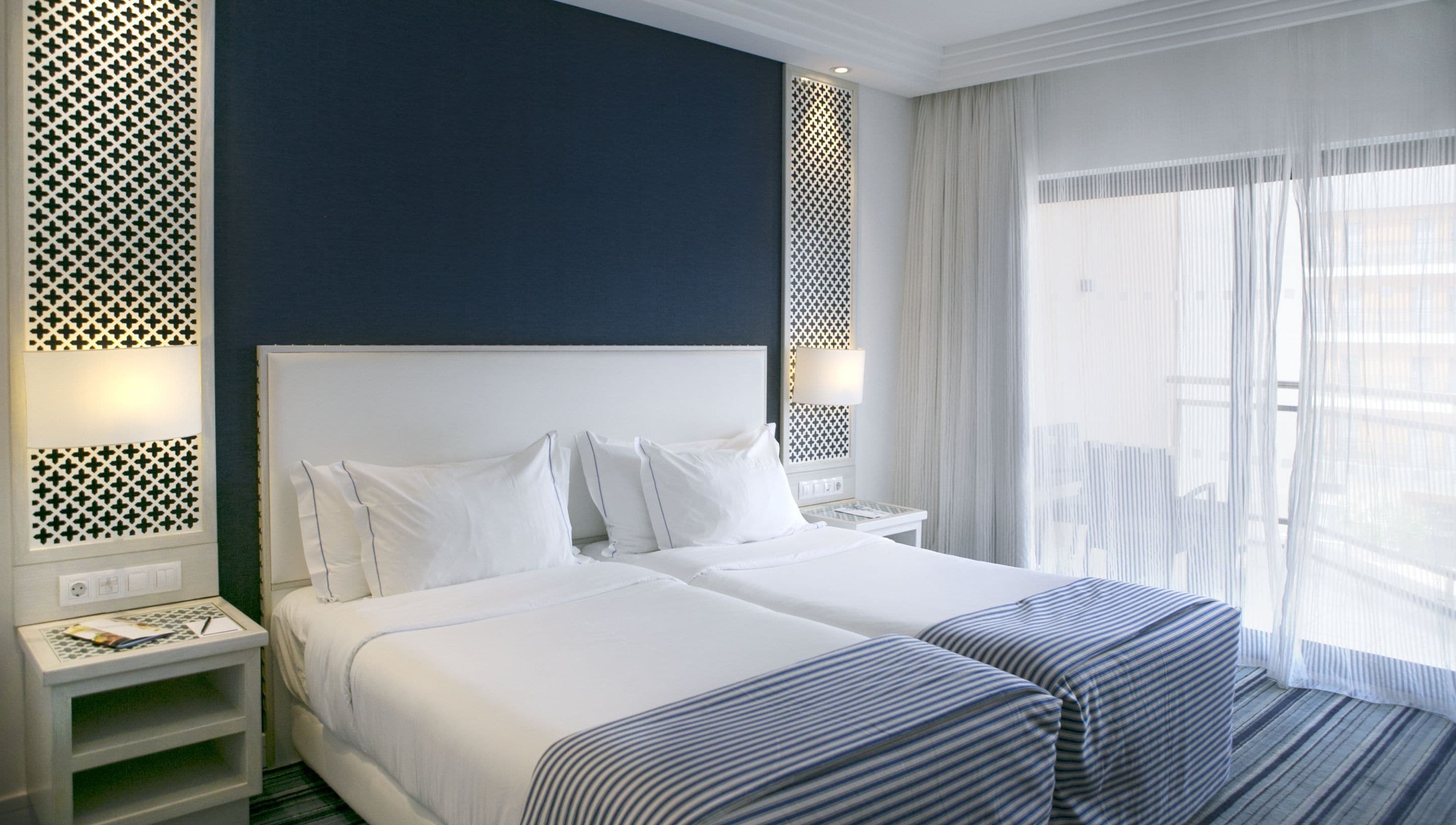 A hotel room with a large bed, two bedside tables, a nightstand, and a window with sheer curtains.