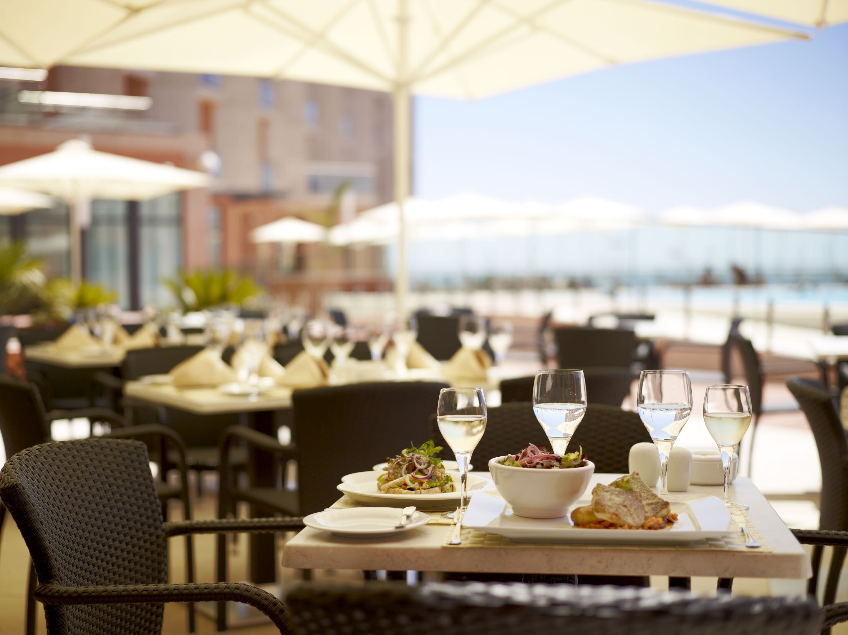 Un área de comedor al aire libre con mesas preparadas para dos personas, con platos blancos, ensaladas en cuencos y copas de vino blanco.
