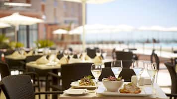 Un área de comedor al aire libre con mesas preparadas para dos personas, con platos blancos, ensaladas en cuencos y copas de vino blanco.