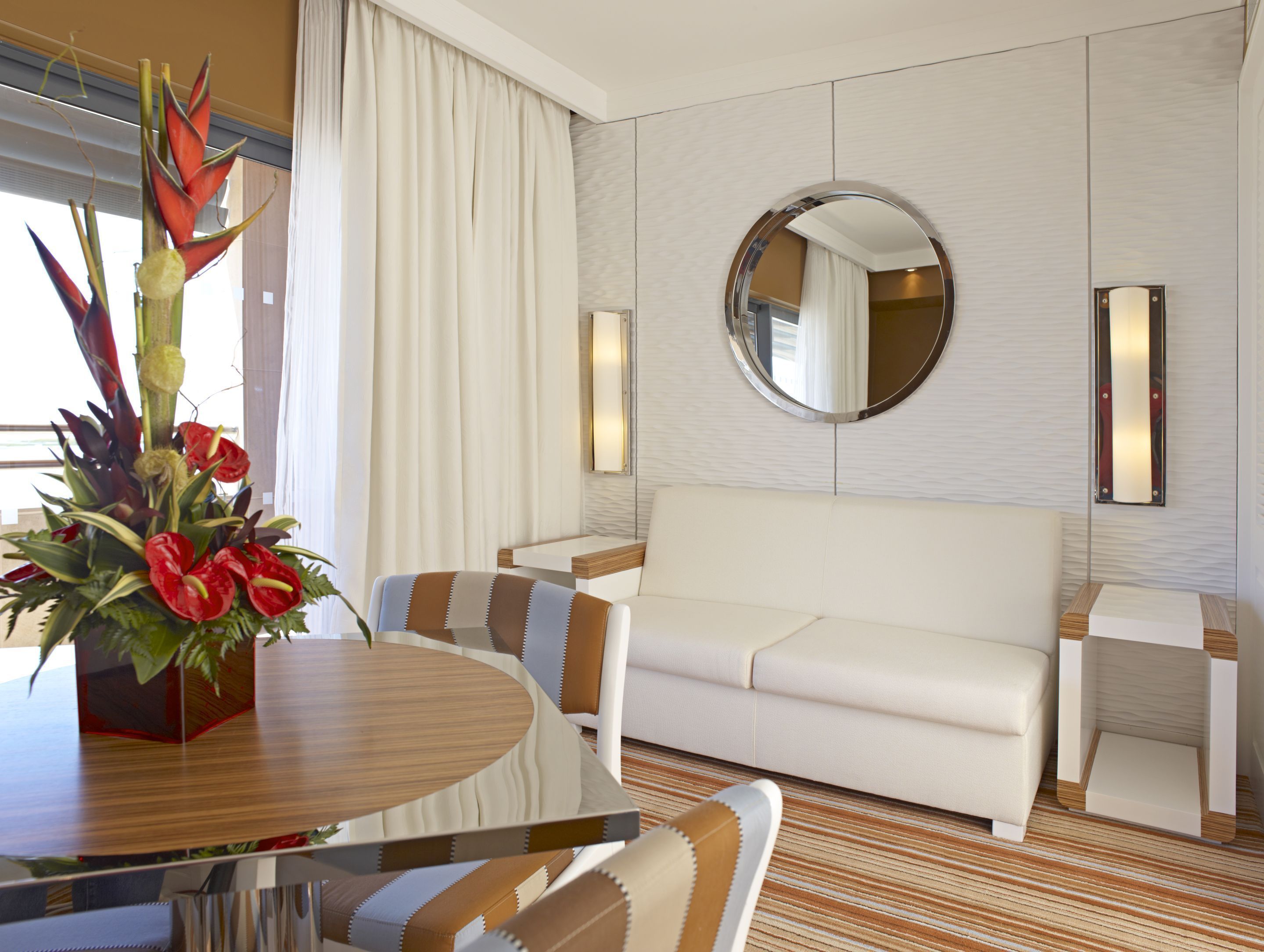 Una habitación de hotel moderna con una mesa de comedor redonda, un sofá blanco y un espejo redondo en la pared.