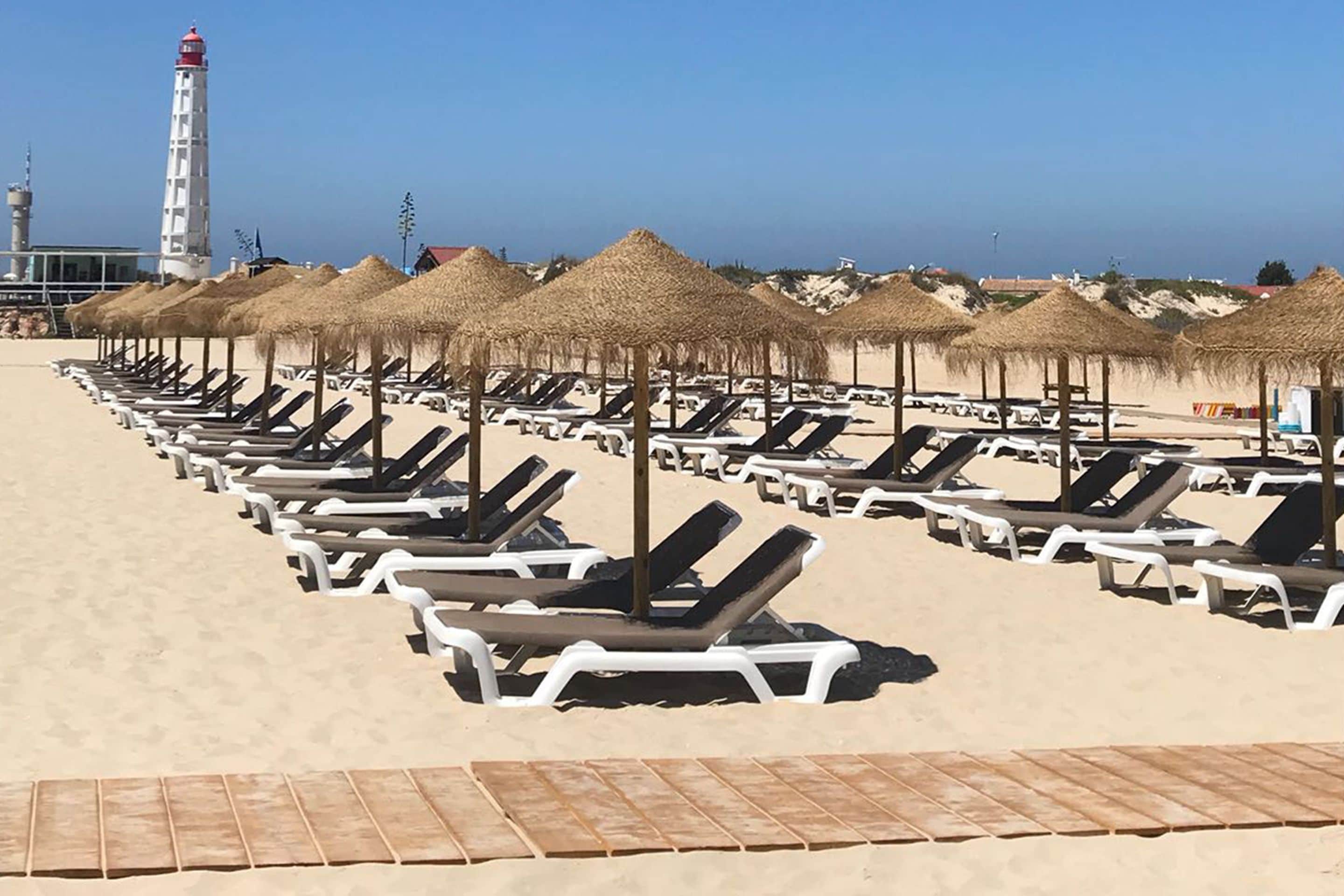 A beach with rows of sun loungers and thatched umbrellas, a lighthouse in the background, and a clear sky.