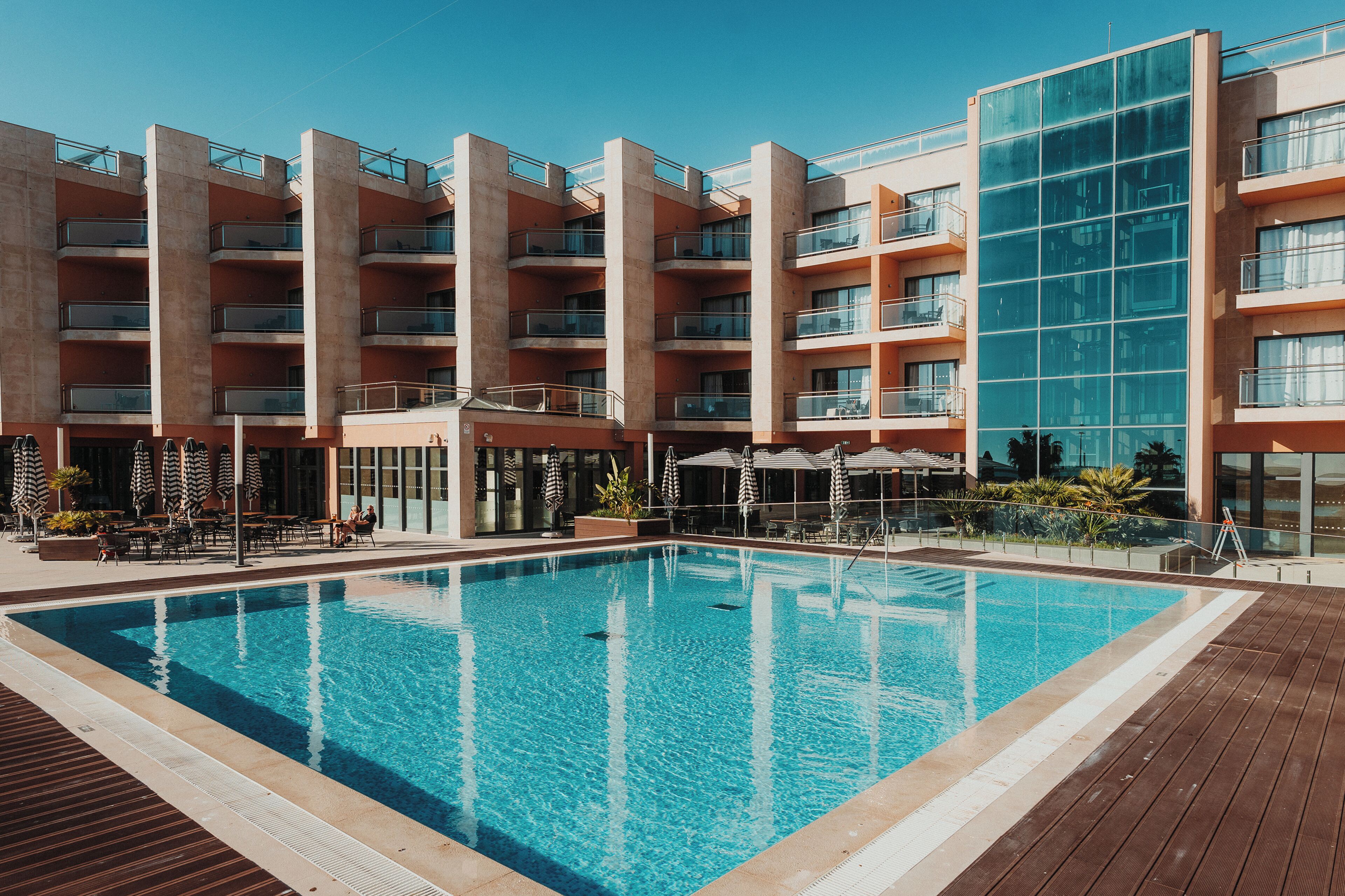 A modern hotel with a large outdoor swimming pool, multiple balconies, and outdoor seating areas.
