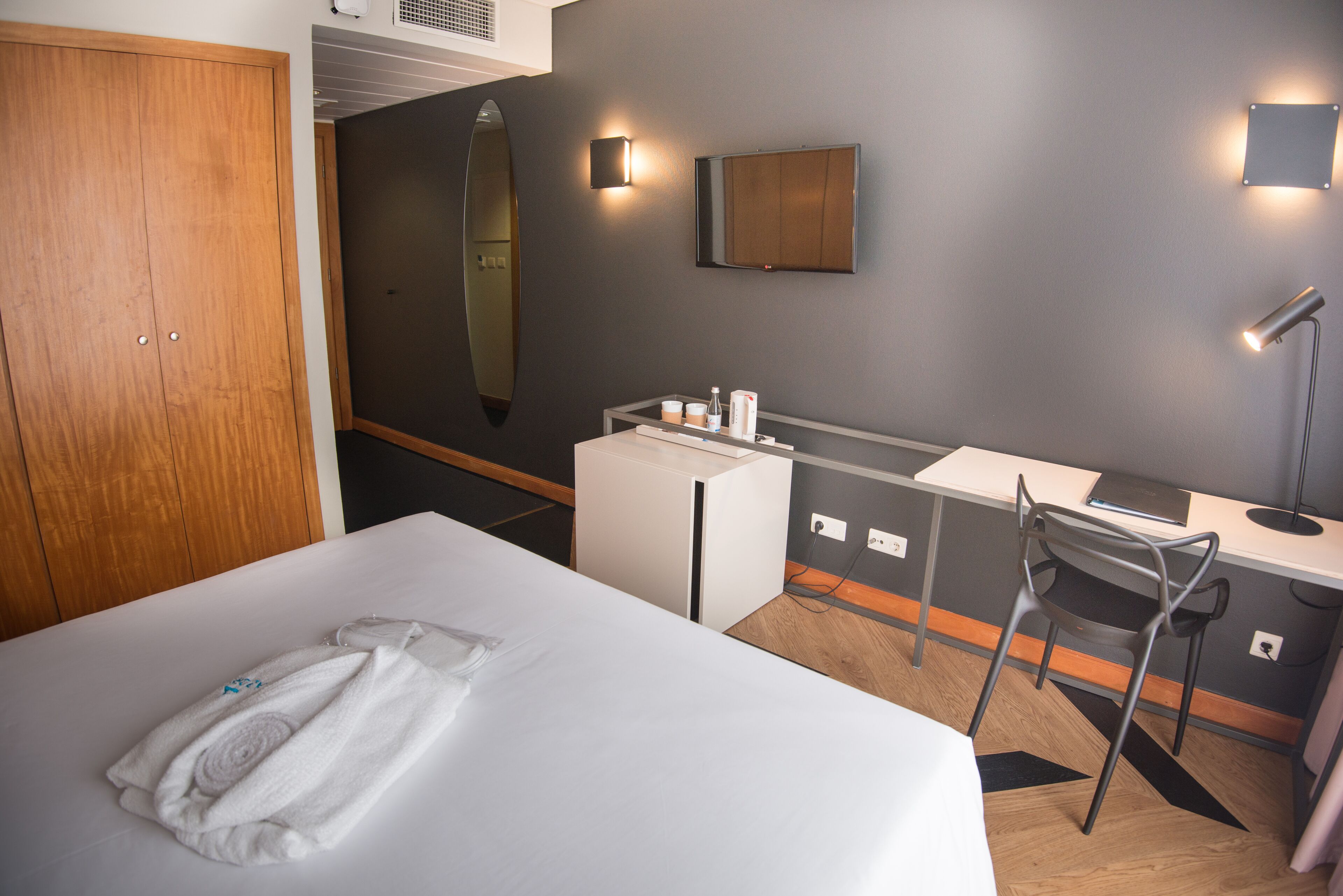A hotel room with a bed, a desk, a chair, a mirror, and a small kitchenette.