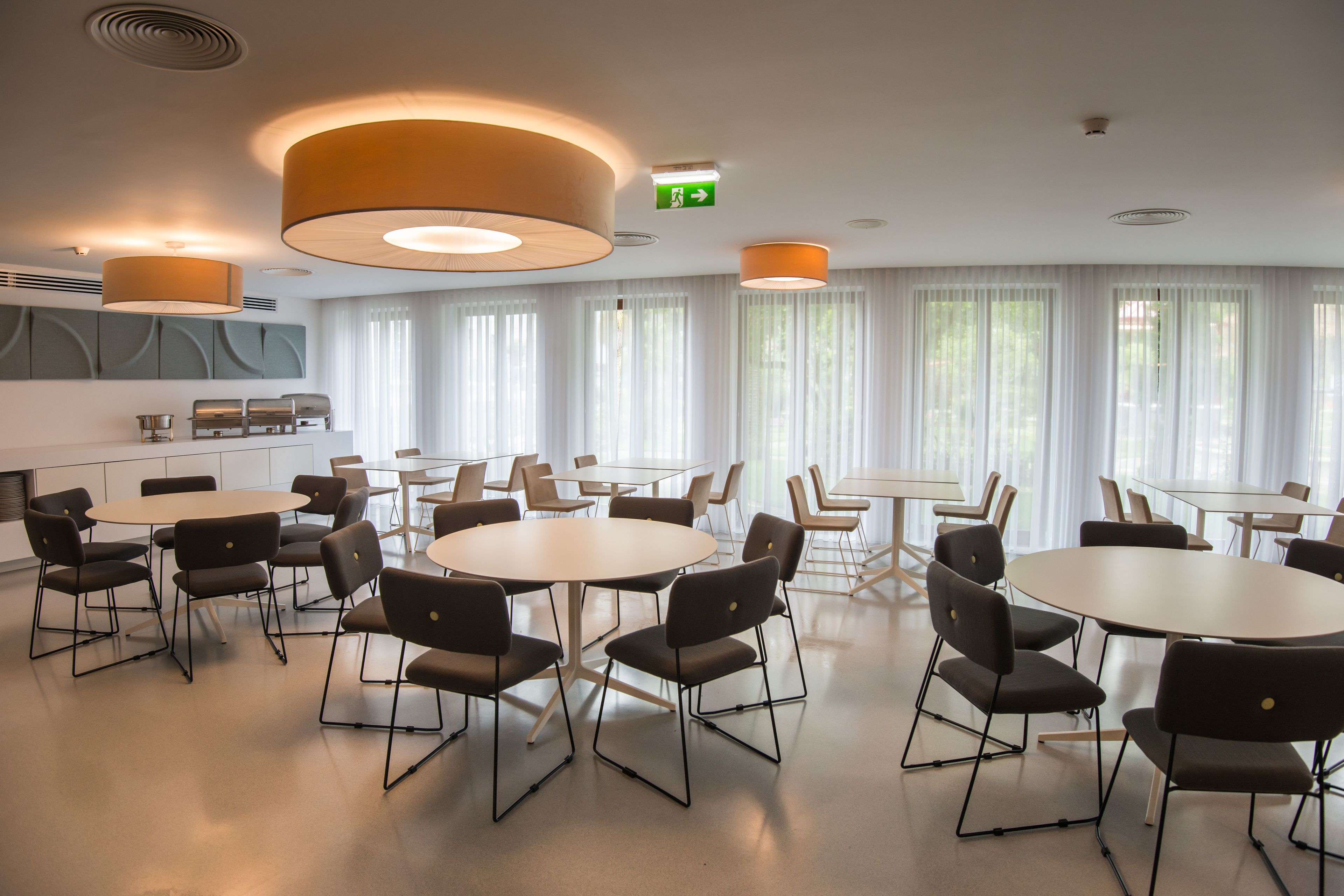 Une salle de conférence moderne, dotée de tables et de chaises rondes, de grandes fenêtres avec des rideaux légers et d’un éclairage encastré.