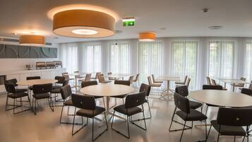 Une salle de conférence moderne, dotée de tables et de chaises rondes, de grandes fenêtres avec des rideaux légers et d’un éclairage encastré.