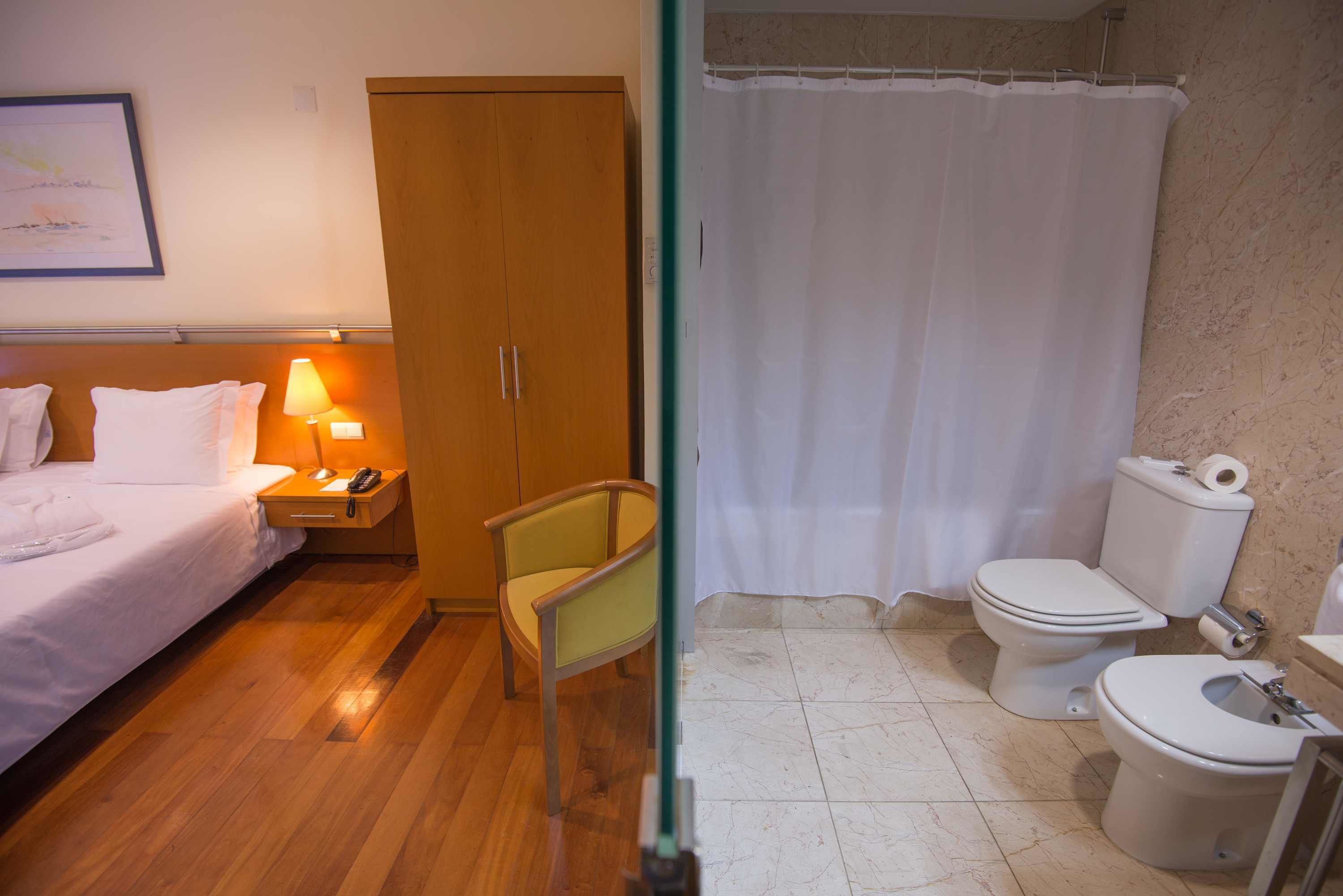 A hotel room with a bed, nightstand, lamp, chair, and a bathroom with a toilet and shower curtain.