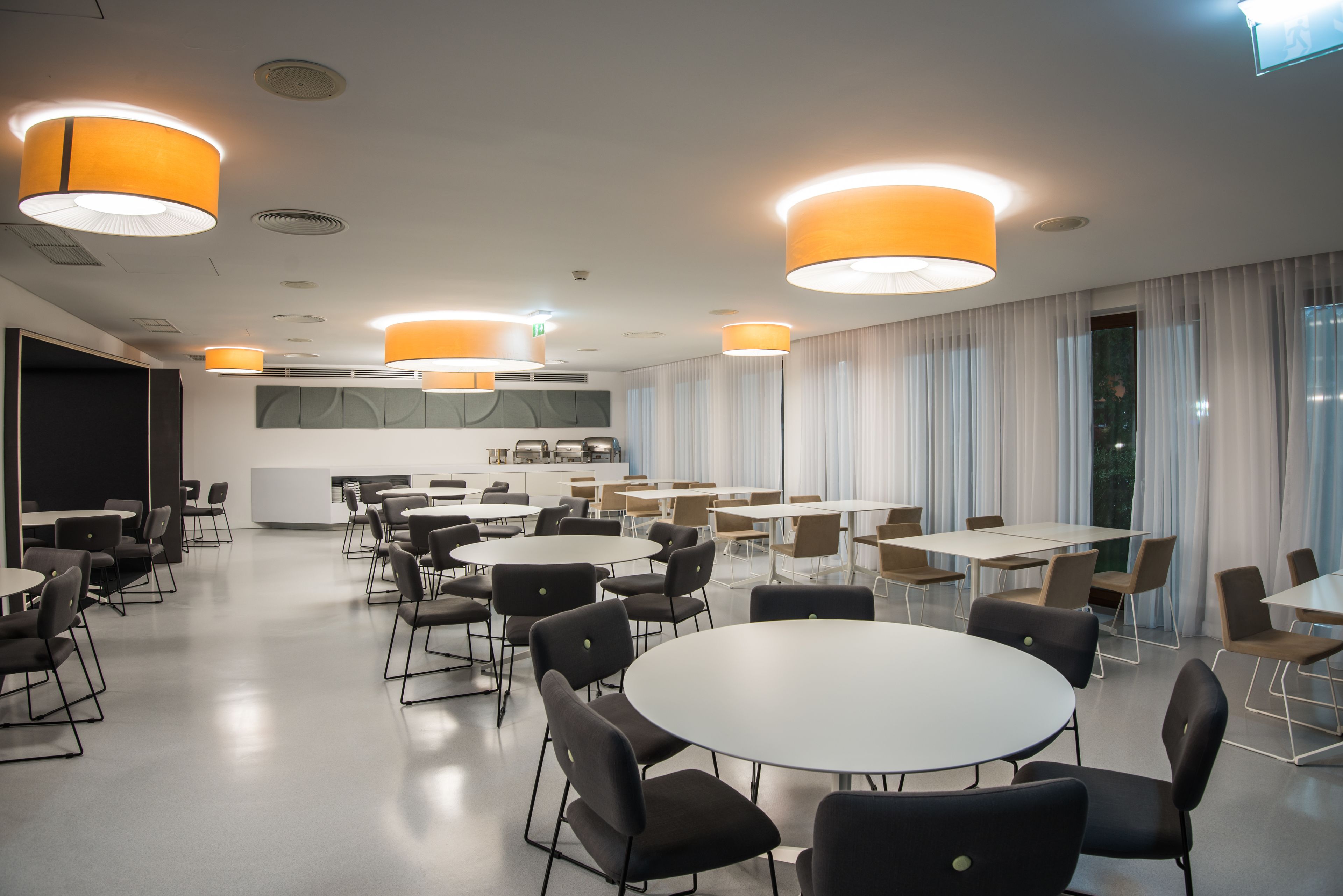 Een moderne vergaderruimte met ronde tafels en stoelen, grote ramen met gordijnen, en een keuken in de achtergrond.