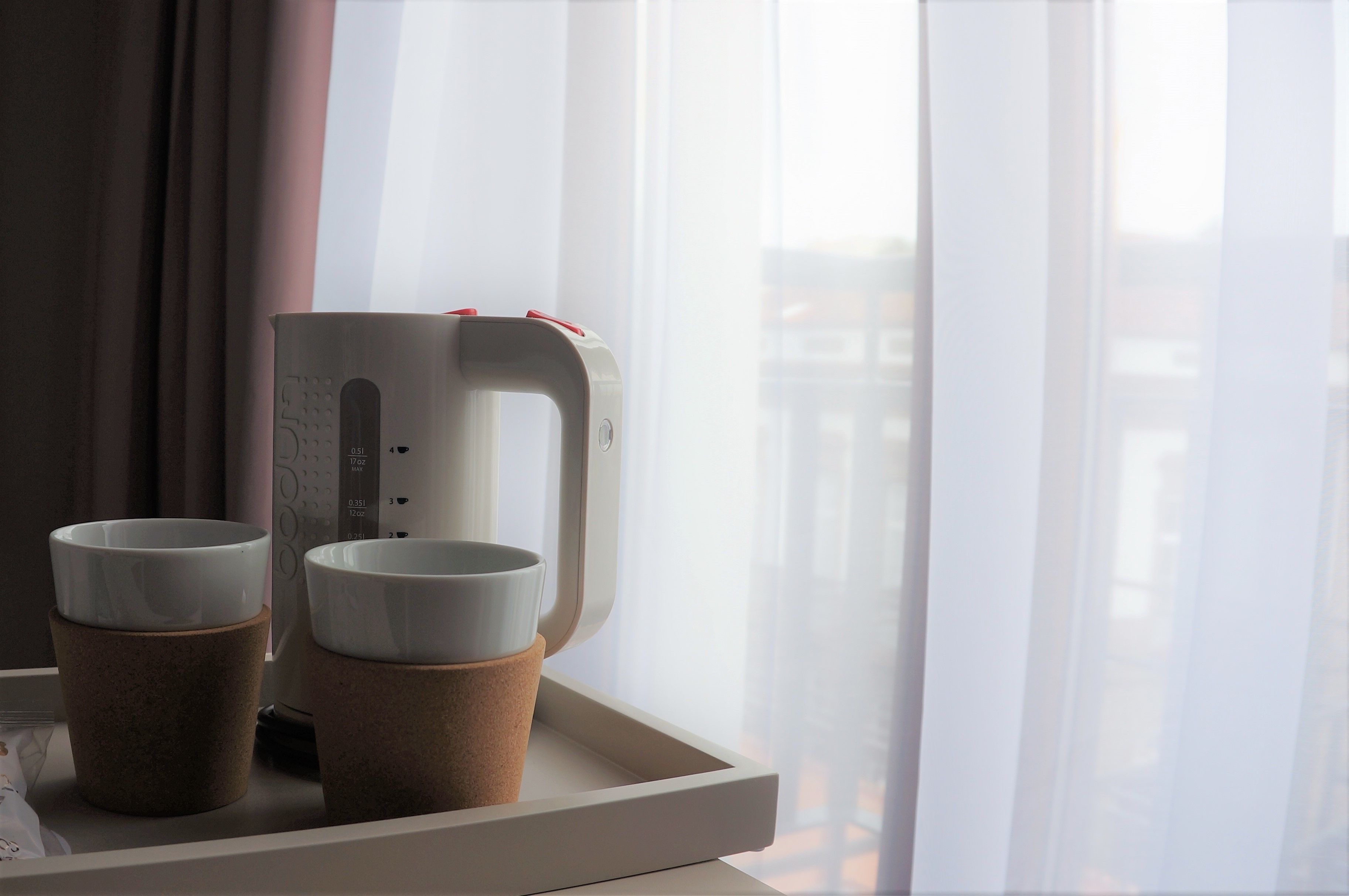 A coffee maker on a tray with two cups, a window with curtains, and a view of buildings.