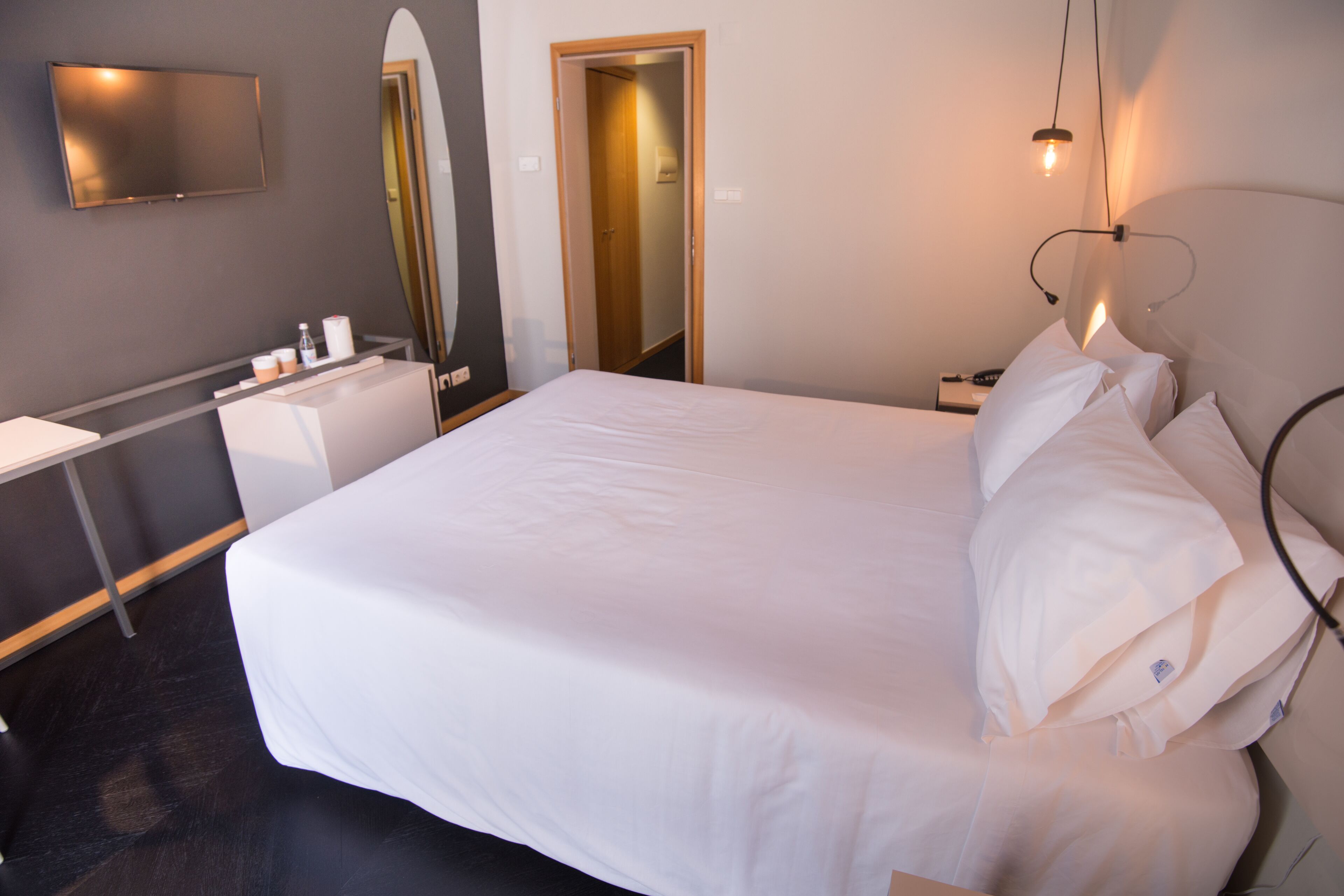 A hotel room with a large bed, a desk with a chair, a flat-screen TV, a mirror, and a bedside table with a lamp.