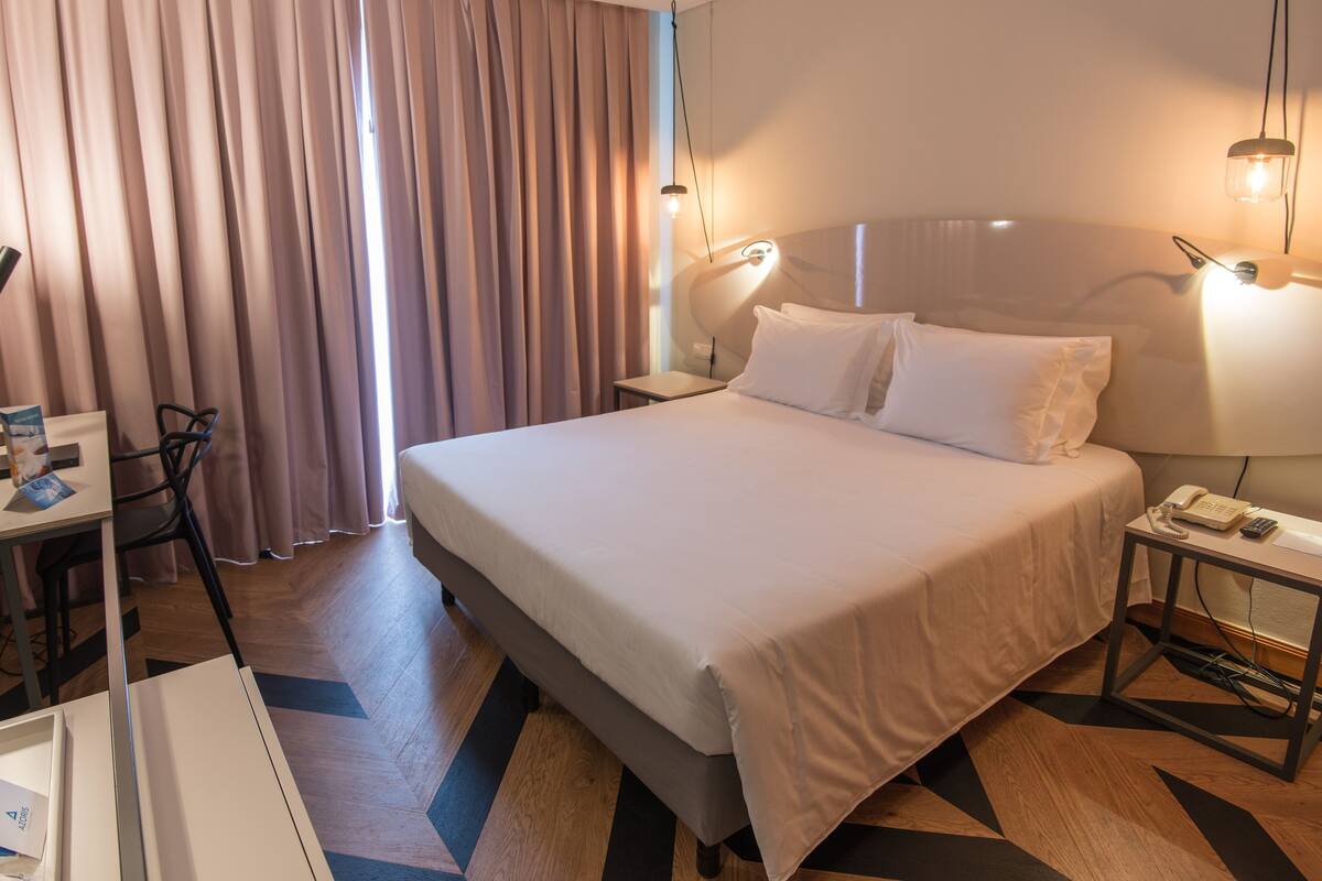 A hotel room with a large bed, a desk with a chair, a bedside table with a lamp, and a window with curtains.