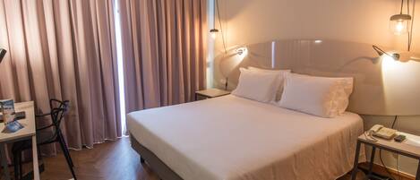 Habitación de hotel con una cama grande, un escritorio con una silla, una mesita de noche con lámpara y una ventana con cortinas.