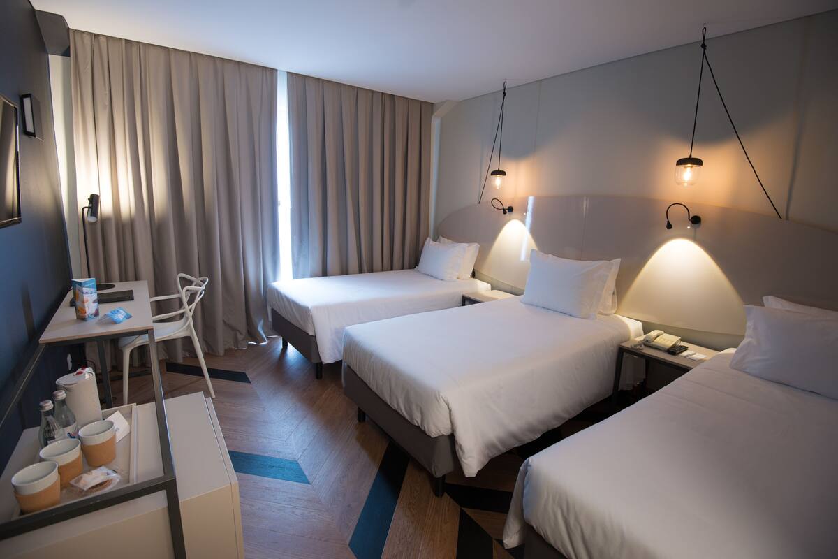 A hotel room with three beds, a desk with a chair, a small table with cups and bottles, and a nightstand with a phone.