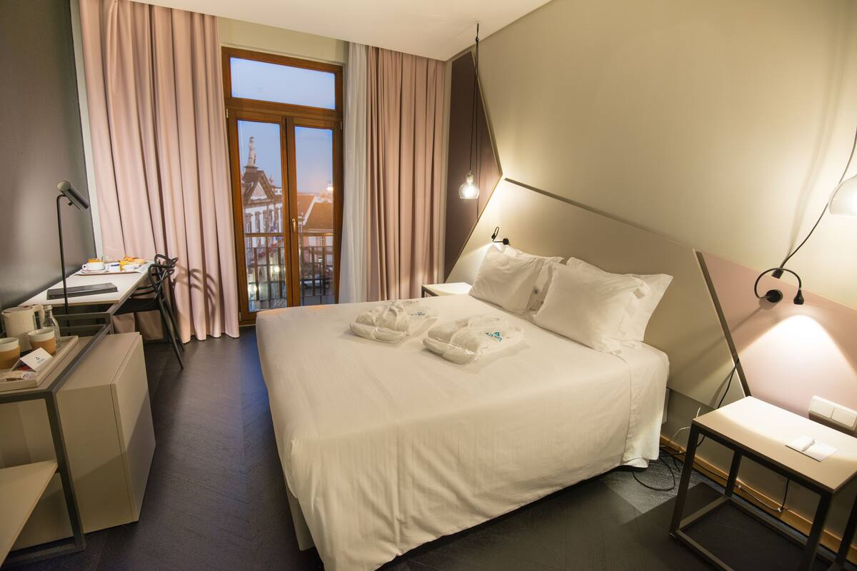 A hotel room with a bed, a desk, a chair, and a view of a building through the window.