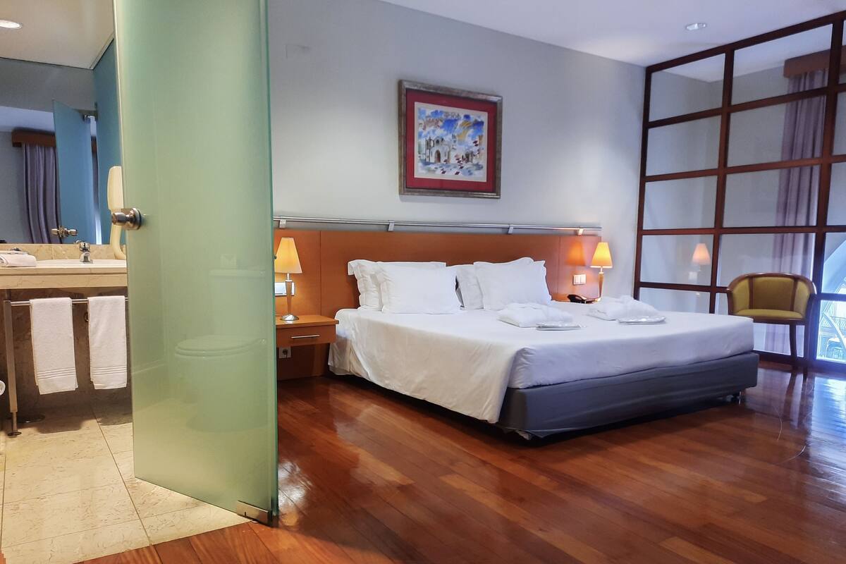 A hotel room with a large bed, a nightstand with lamps, a bathroom with a sink and towel rack, and a window with a view.