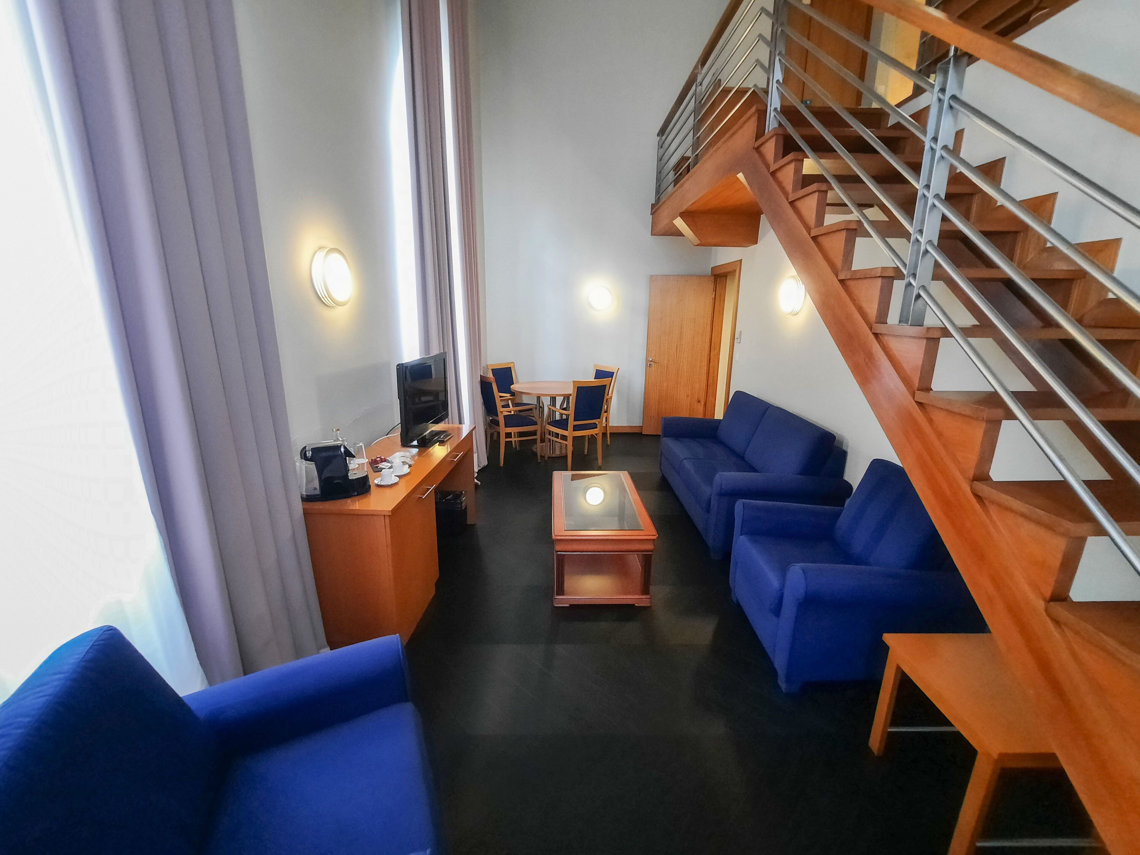 A hotel room with a wooden staircase, blue sofas, a wooden desk with a TV, and a dining area.