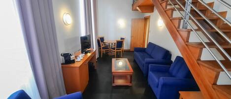A hotel room with a wooden staircase, blue sofas, a wooden desk with a TV, and a dining area.