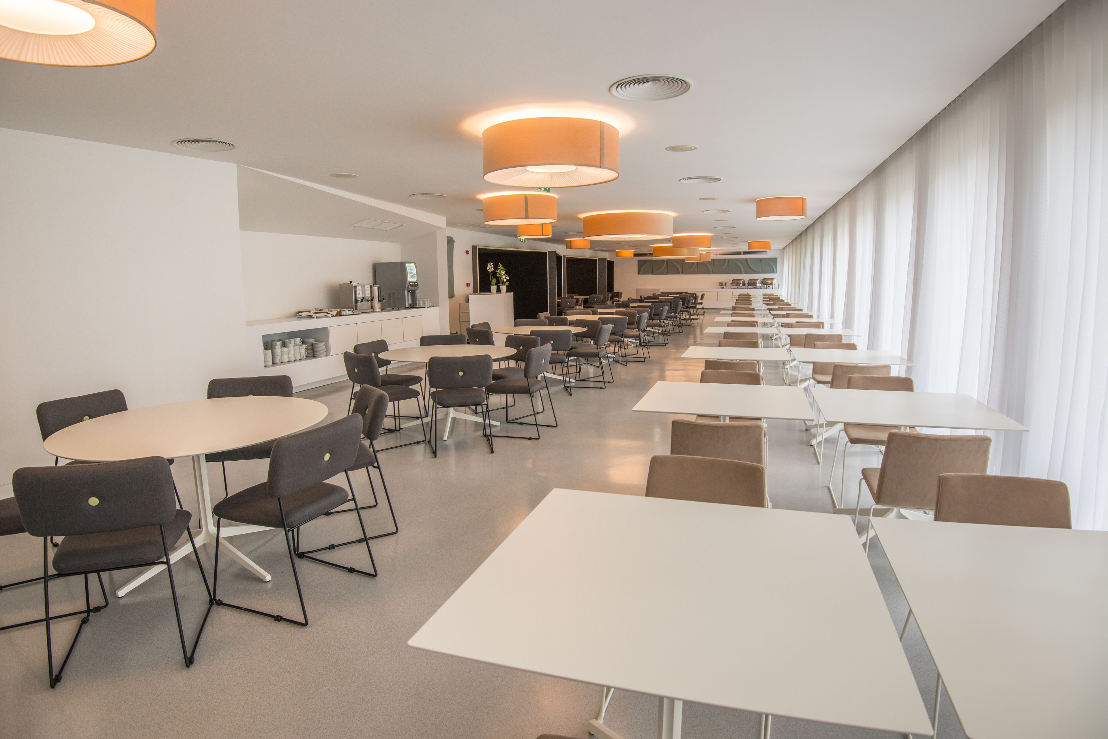 A modern dining area with round and square tables, black chairs, and orange pendant lights.
