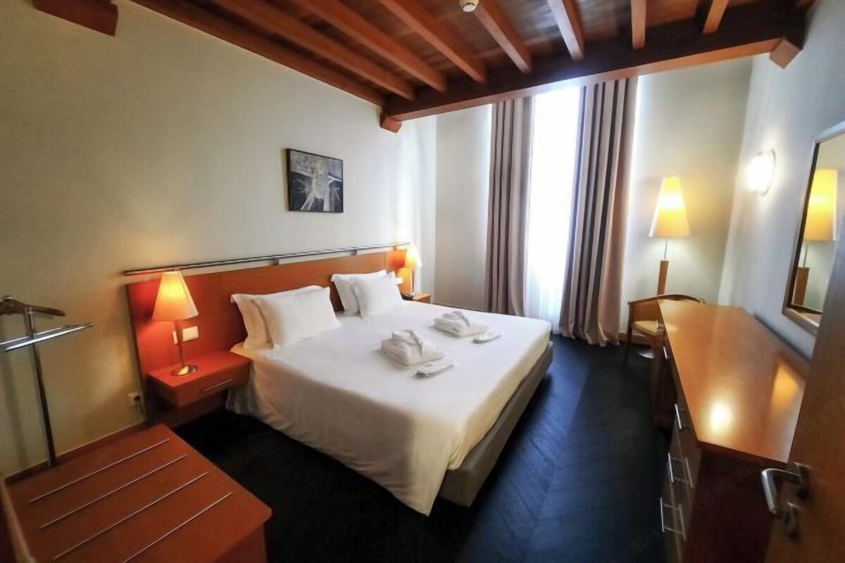Une chambre d’hôtel comprenant un lit, des lampes de chevet, une tête de lit en bois, une table de chevet, un bureau et un miroir.
