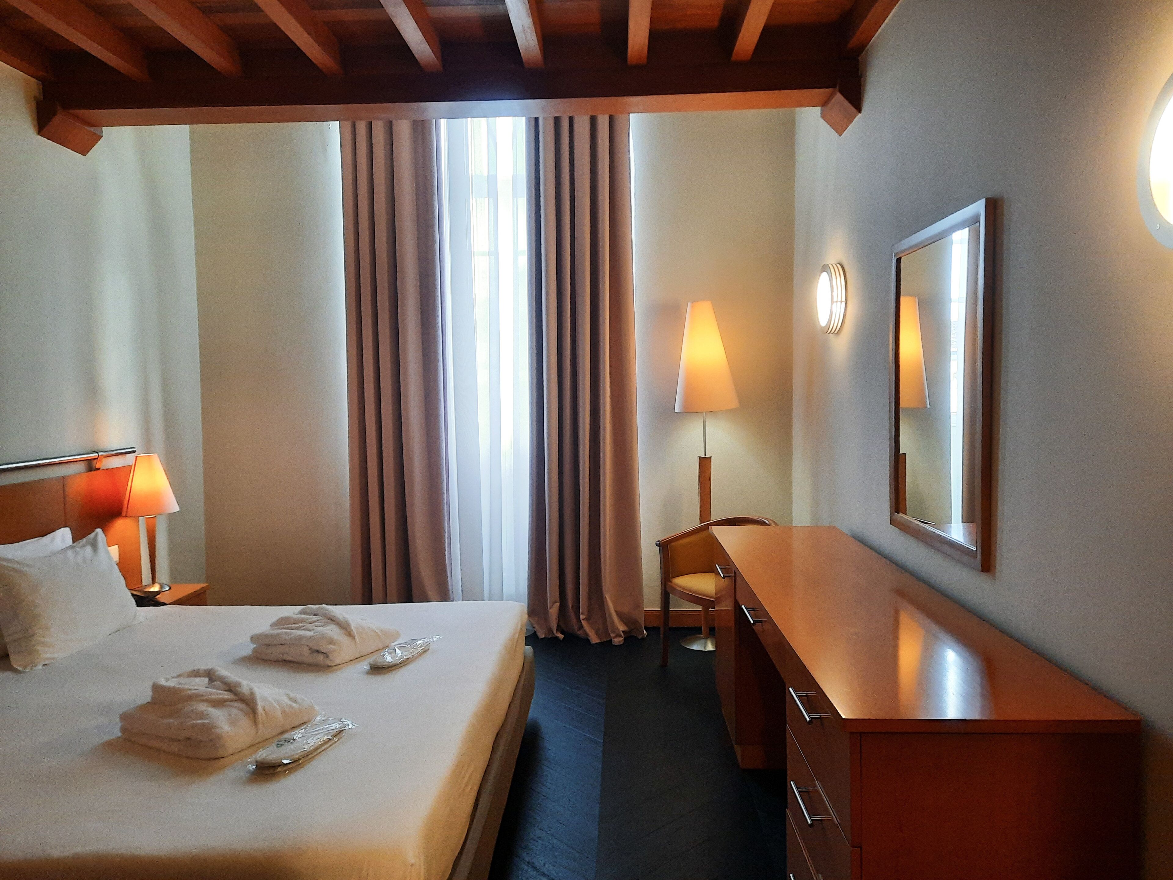 A hotel room with a bed, a desk, a chair, a lamp, and a mirror.