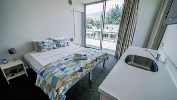 A hotel room with a bed, a small bedside table with a lamp, a sink, and a large window with a view of buildings and trees.