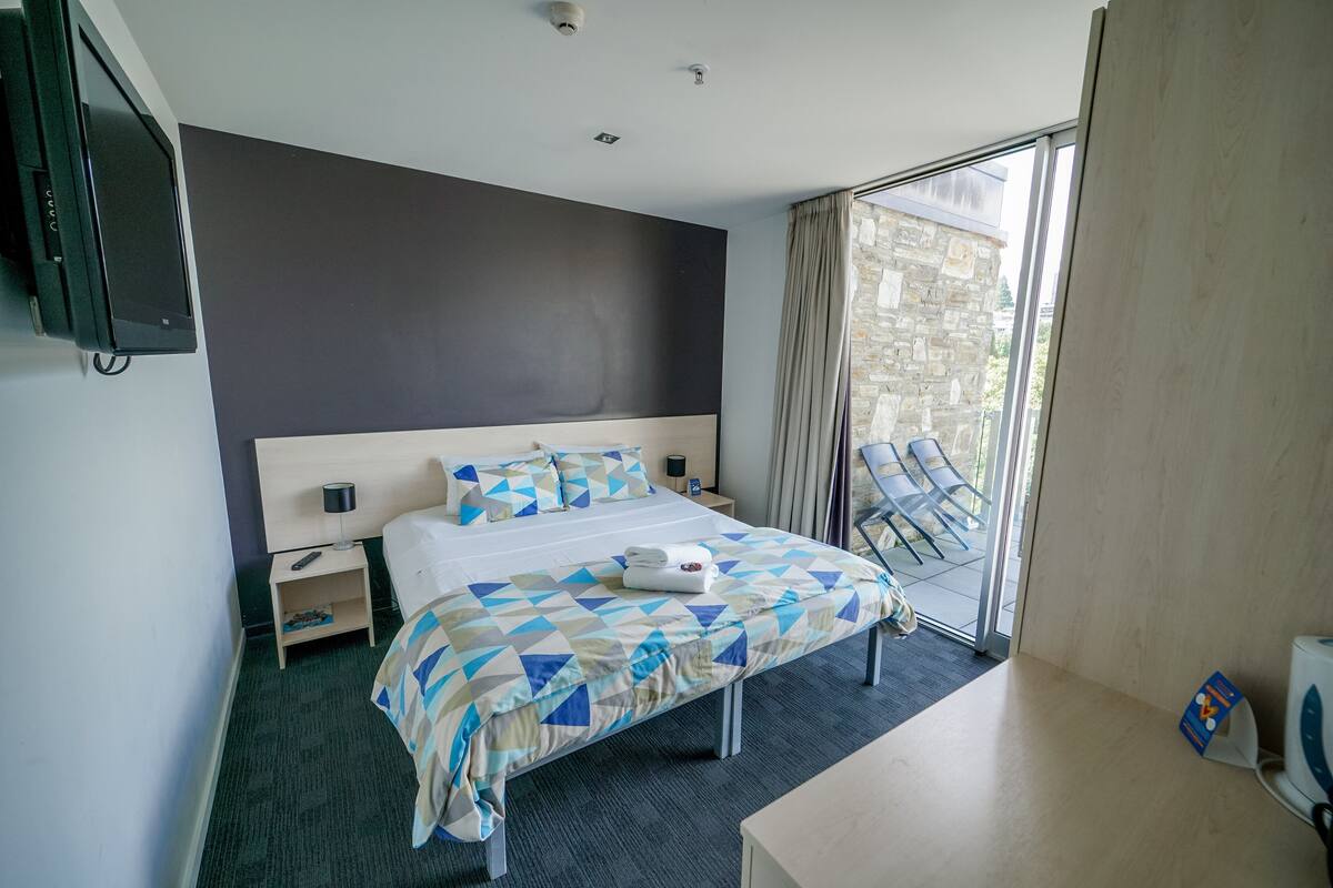 A hotel room with a large bed, a desk, a TV, and a balcony with chairs.