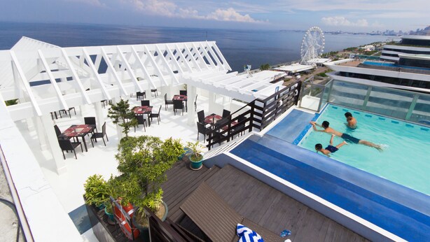 A rooftop pool area with a view of the ocean, outdoor seating, and a Ferris wheel in the background.