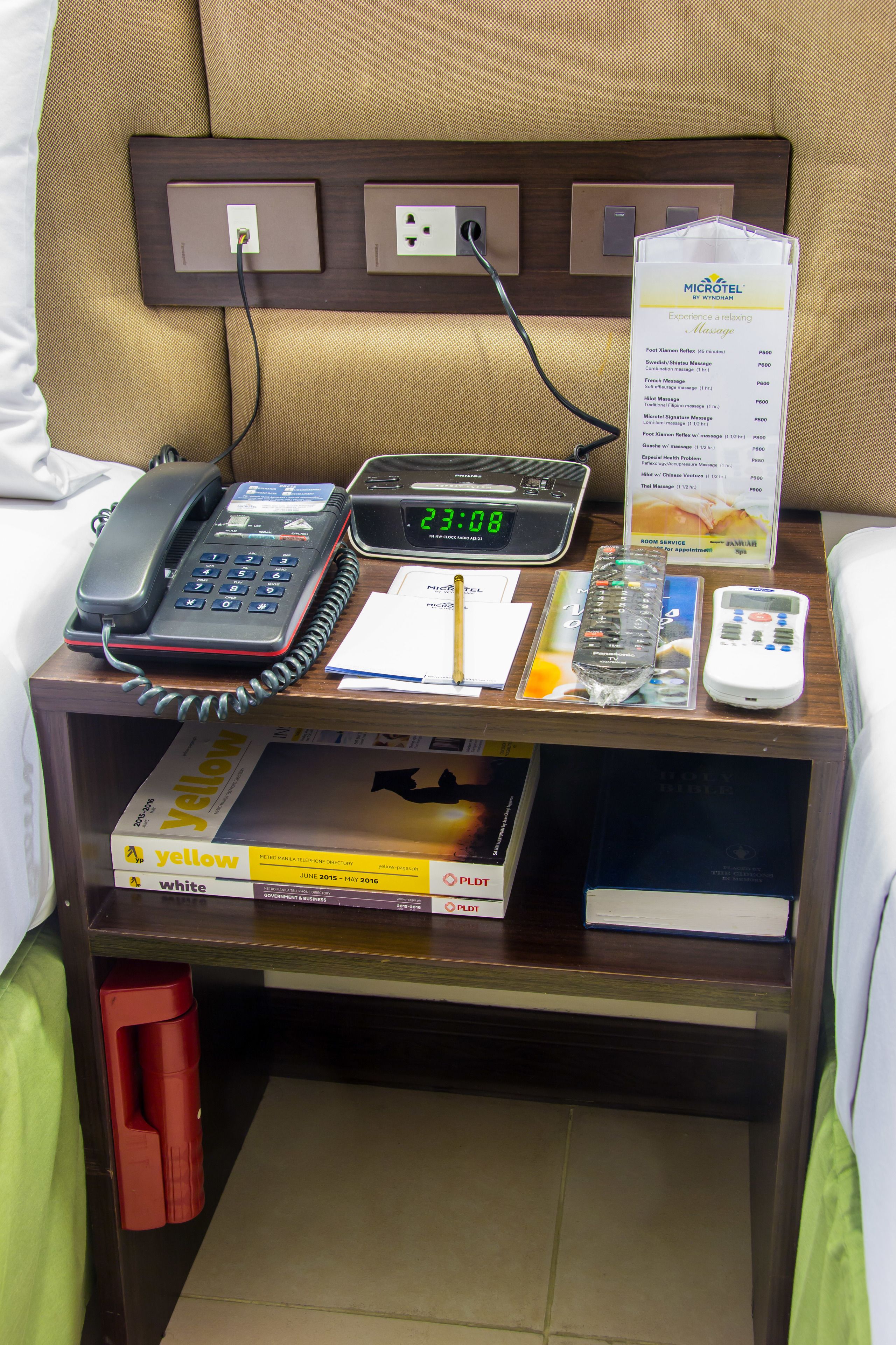 A hotel room with a bedside table containing a phone, a digital clock, a menu, and a remote control.