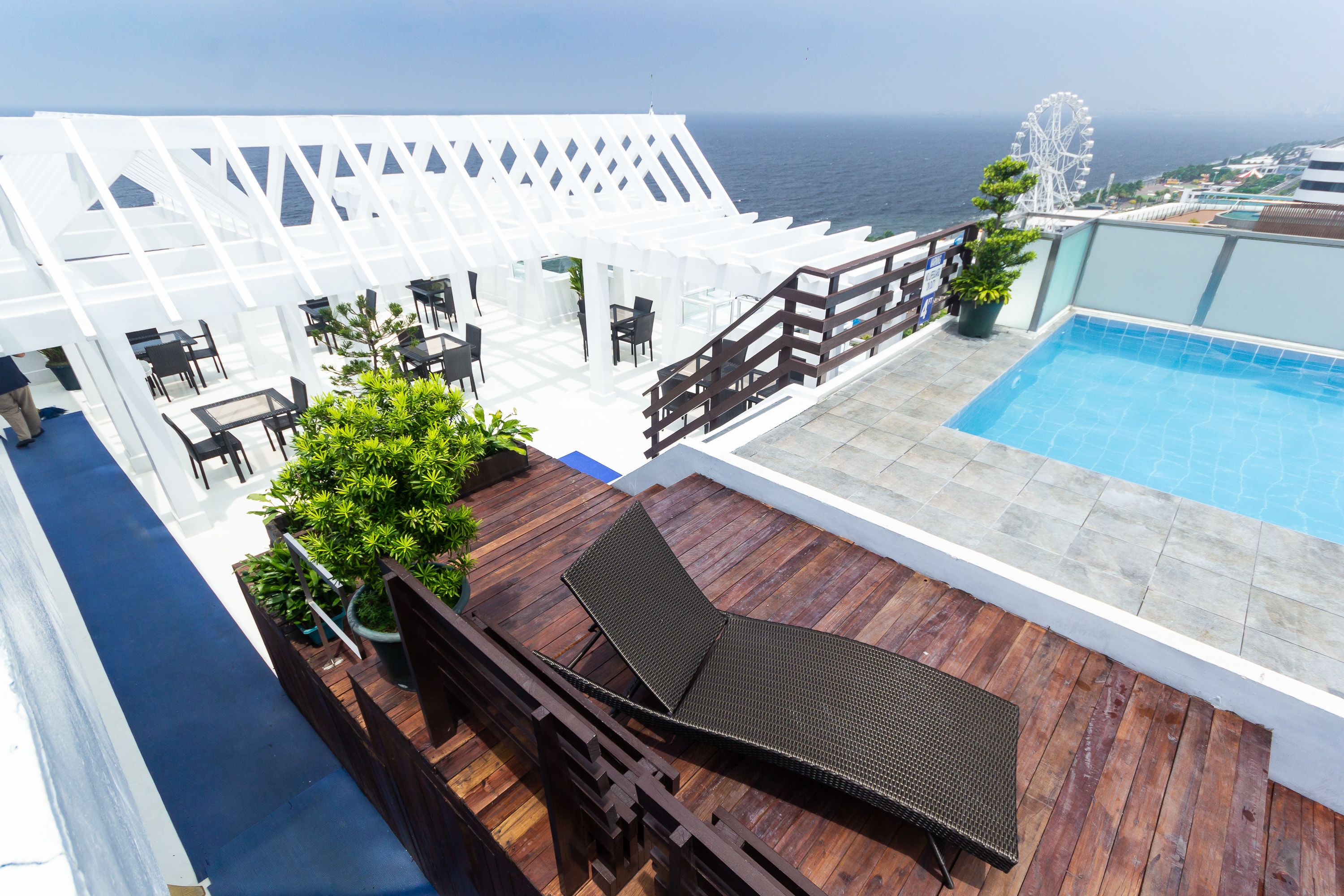A rooftop pool area with a wooden deck, lounge chairs, and a view of the ocean.