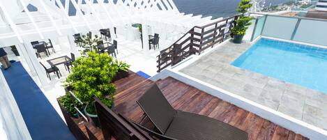 A rooftop pool area with a wooden deck, lounge chairs, and a view of the ocean.