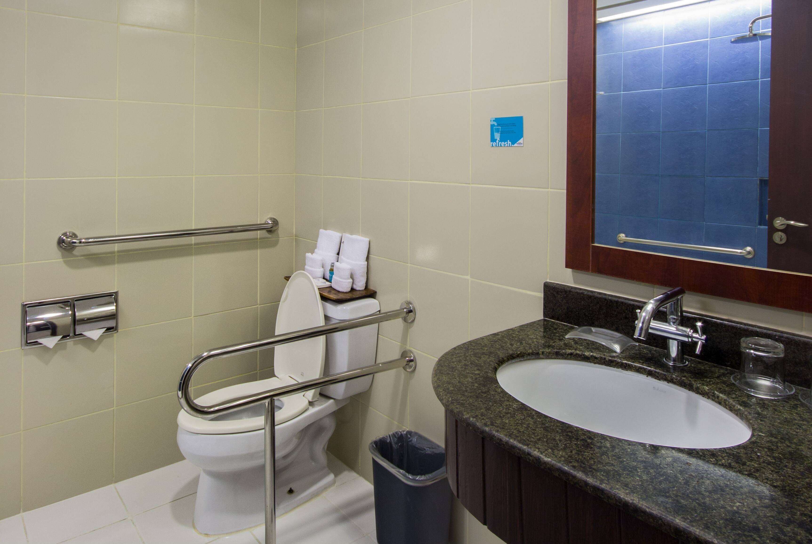 Baño con inodoro, barras de apoyo, lavamanos y cubo de basura.