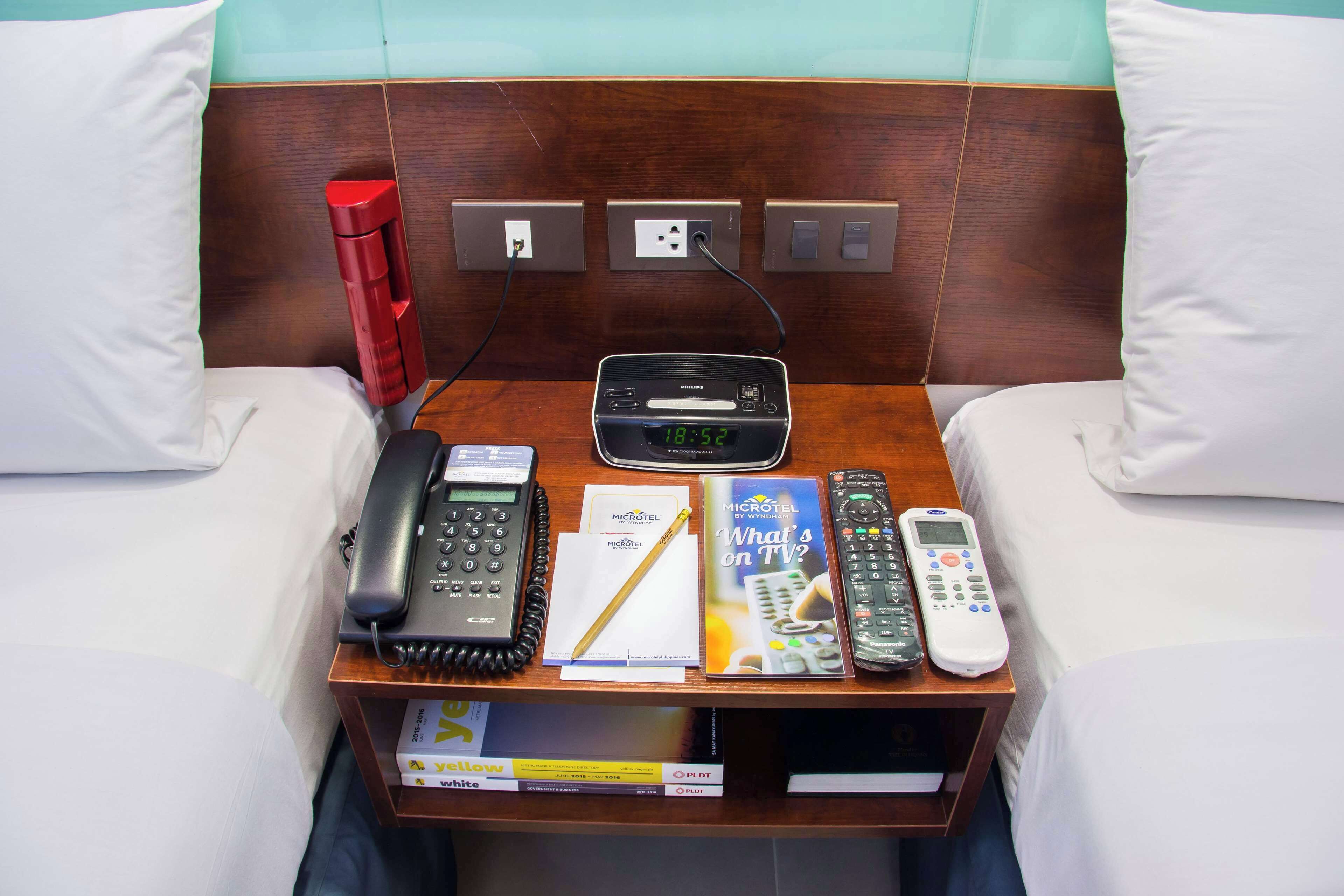 Une chambre d’hôtel avec une tête de lit en bois, une table de chevet sur laquelle se trouve une radio Philips, un téléphone, une télécommande, un magazine et une bouteille d’eau rouge.