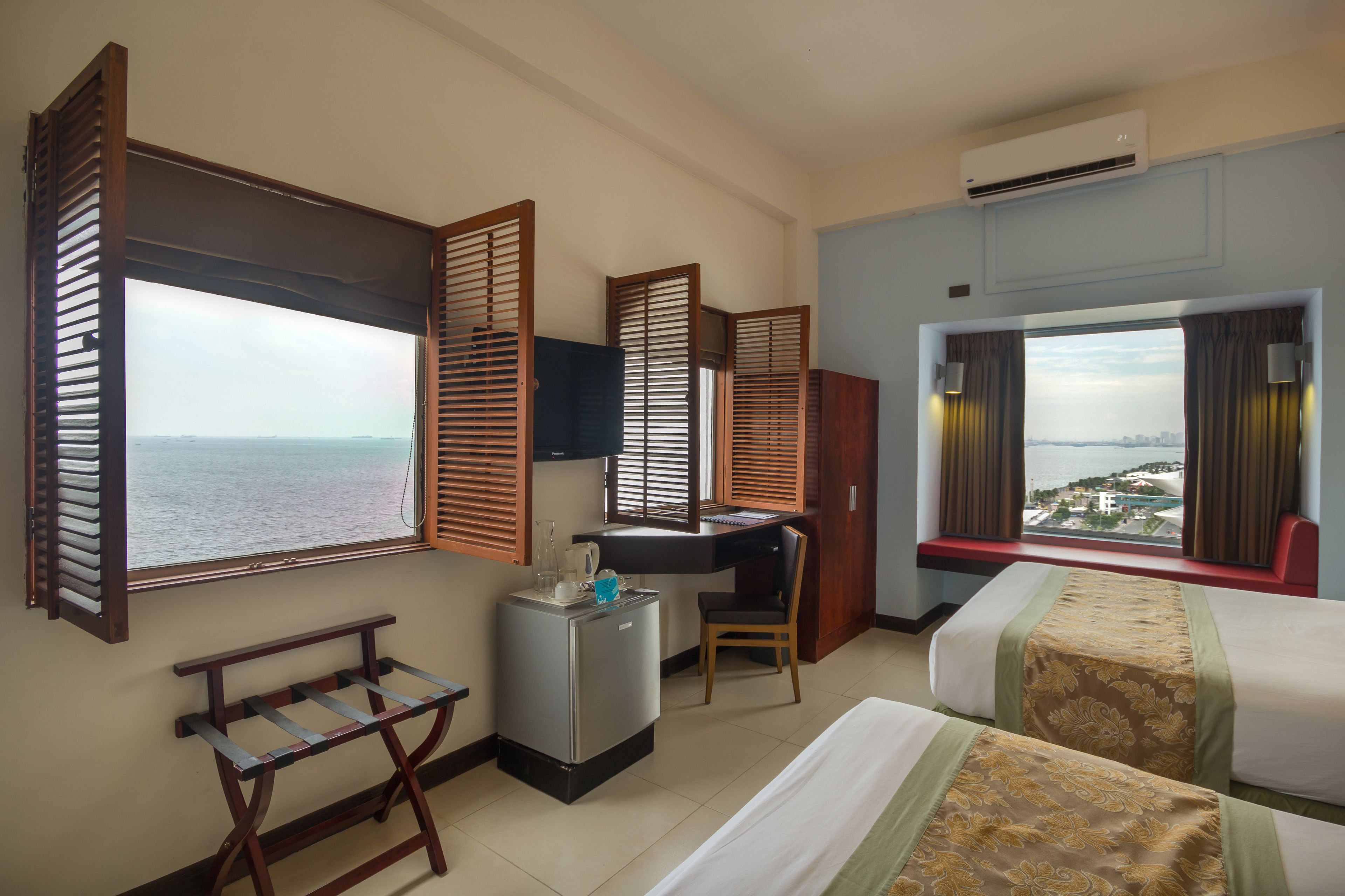 A hotel room with a desk, two beds, a small refrigerator, a bench, and large windows with wooden shutters.