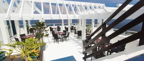 An outdoor terrace with tables and chairs, a white pergola, and a view of the ocean.