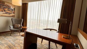 A hotel room with a large window, a desk, a chair, a lamp, and a painting on the wall.