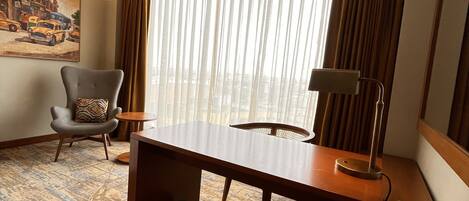 A hotel room with a large window, a desk, a chair, a lamp, and a painting on the wall.