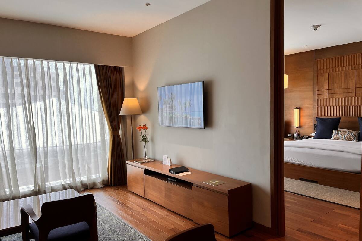 A hotel room with a wooden desk, a chair, a bedside table with a lamp, a TV, and a bed with pillows.