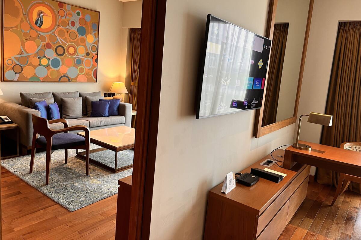 A hotel room with a sofa, armchair, coffee table, TV, and a desk with a lamp.