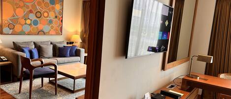 A hotel room with a sofa, armchair, coffee table, TV, and a desk with a lamp.