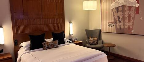 A hotel room with a large bed, a wooden headboard, a chair, a small table, and a wall-mounted artwork.