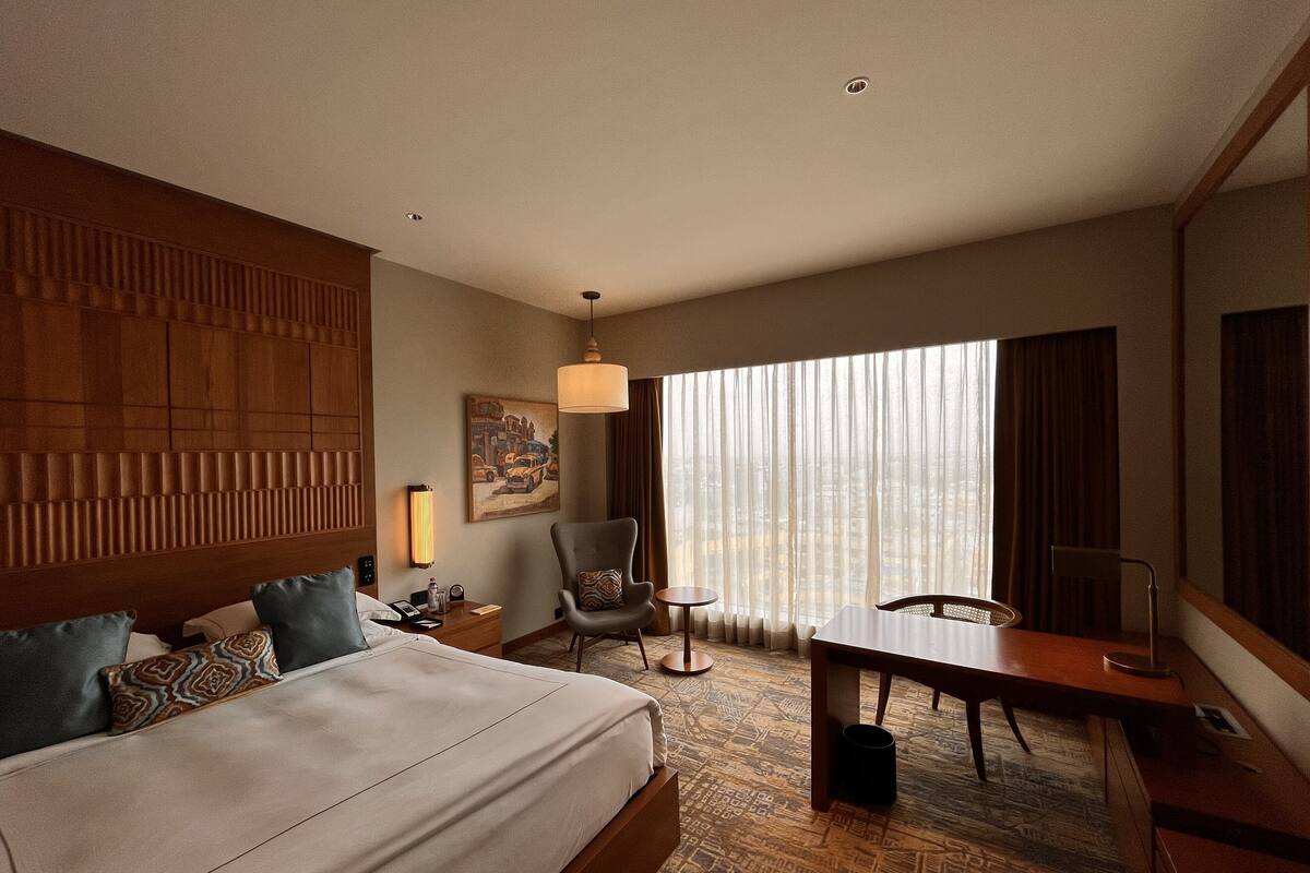 A hotel room with a large bed, a desk, a chair, a lamp, and a view of the city through the window.