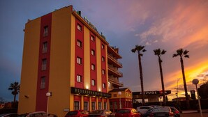 A multi-story hotel with a red and yellow facade, a restaurant on the ground floor, and palm trees in the background. - Hotel Zeus (Merida)