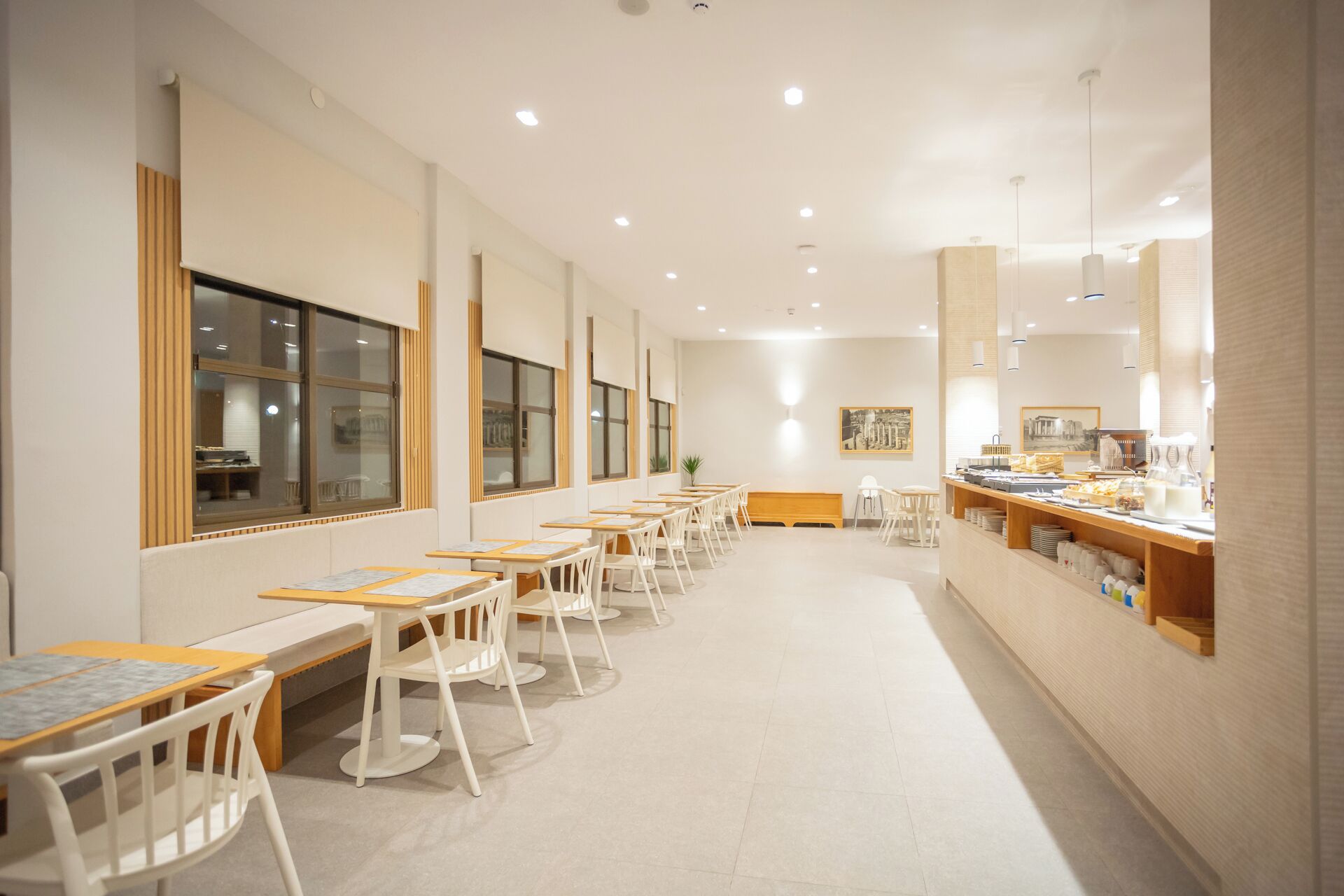 Un café amplio con varias mesas y sillas, una barra con comida y bebidas, y grandes ventanales a lo largo de una pared.