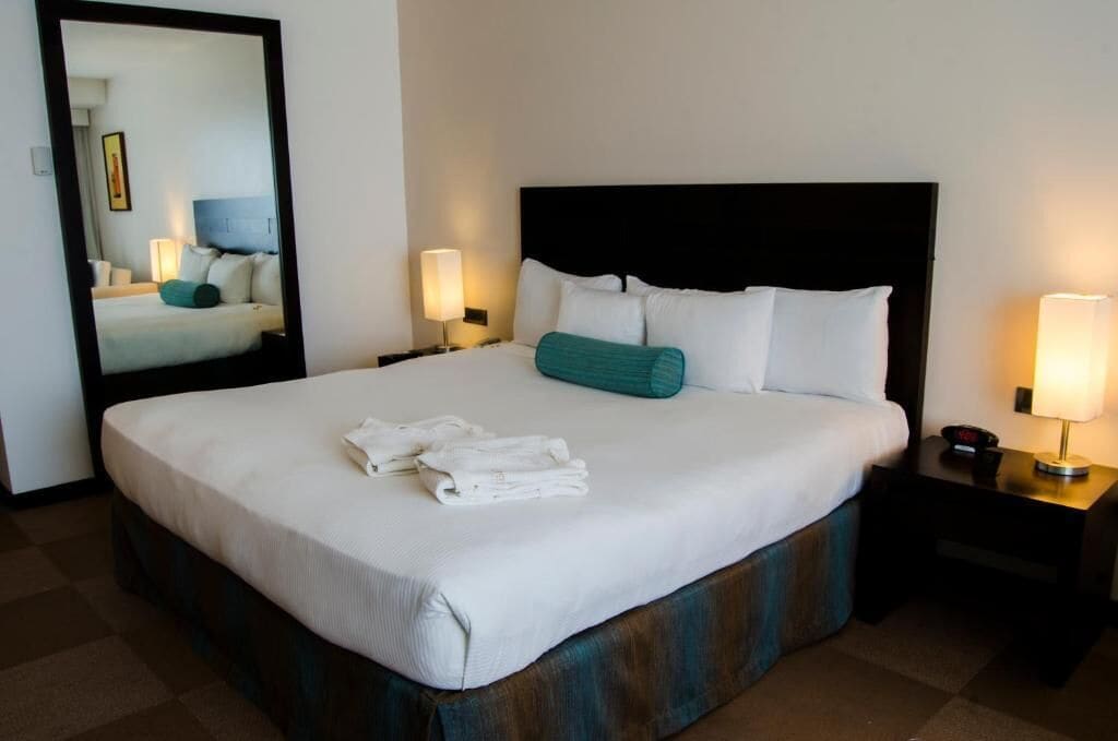 A neatly made bed with white linens and a teal pillow, two bedside lamps, and a nightstand with a clock.