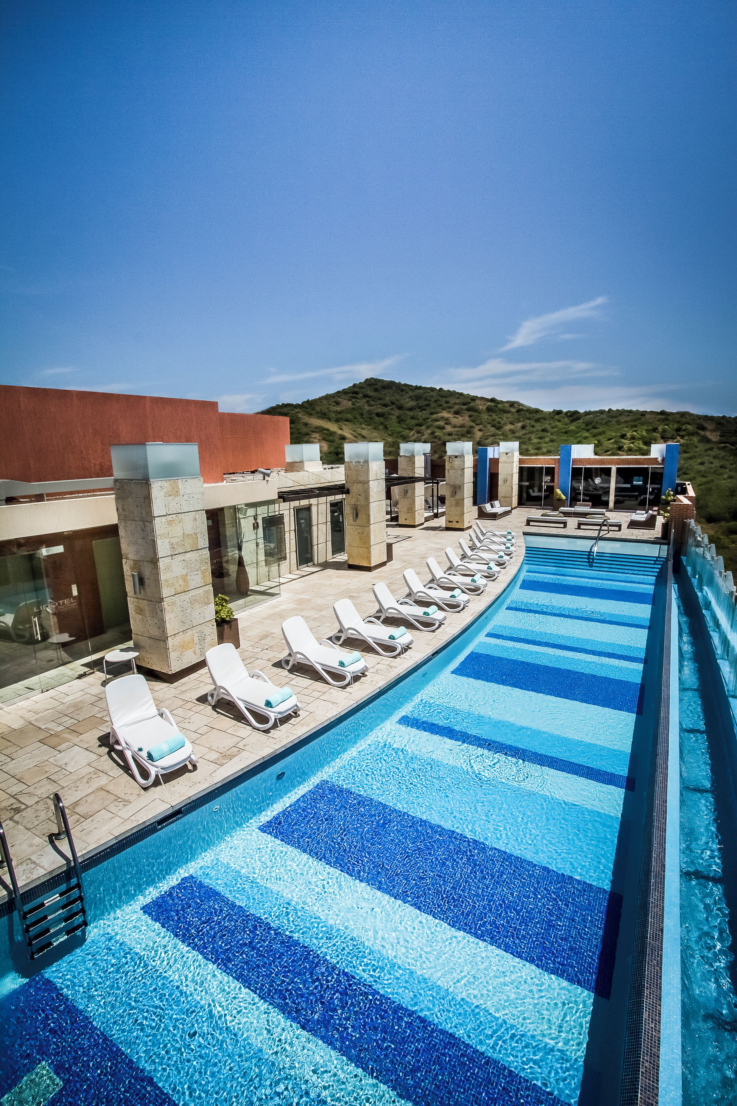 A swimming pool with blue tiles, lounge chairs, and a building with large windows.