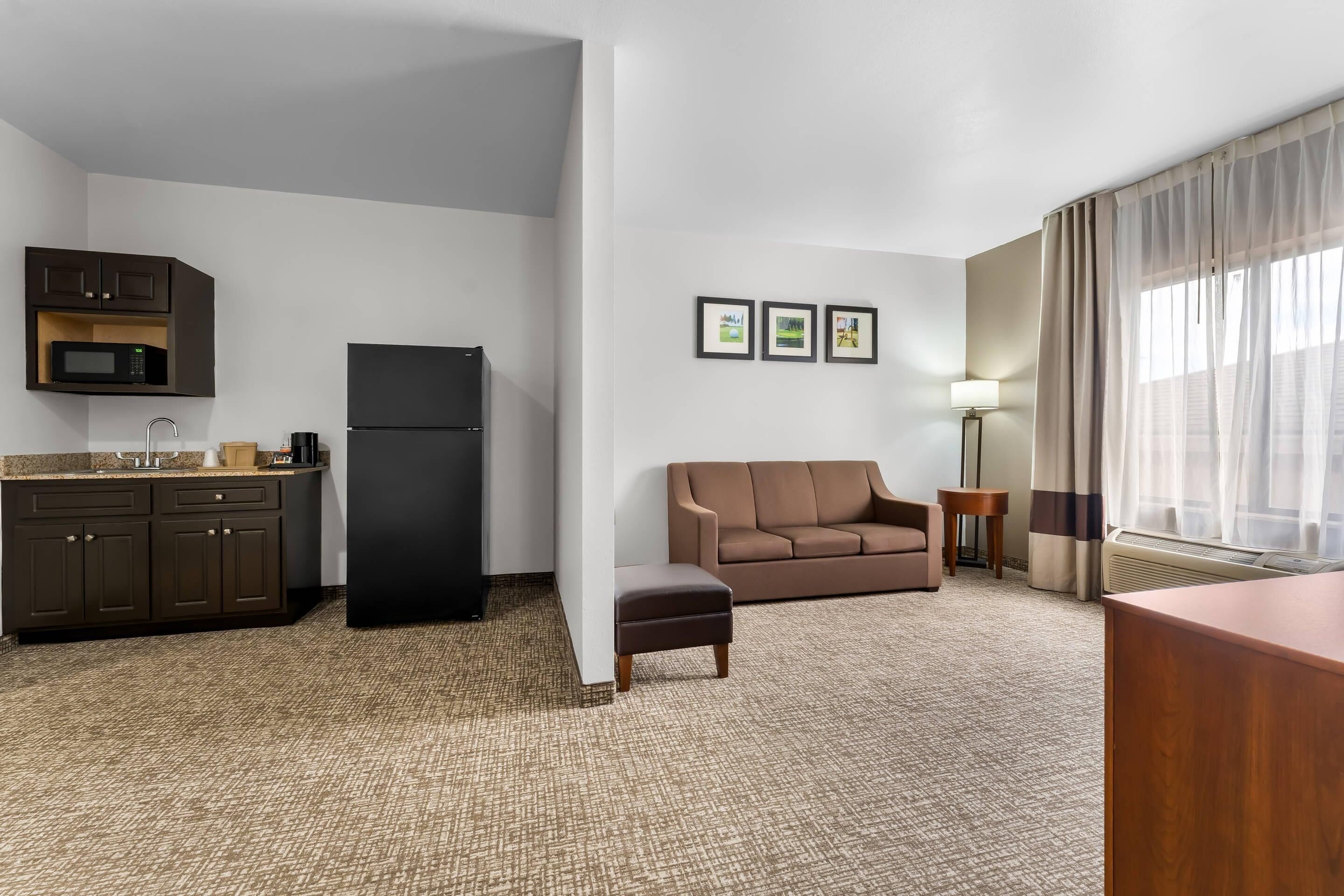 Una habitación de hotel compacta con kitchenette, un sofá marrón, un pequeño reposapiés y una lámpara.