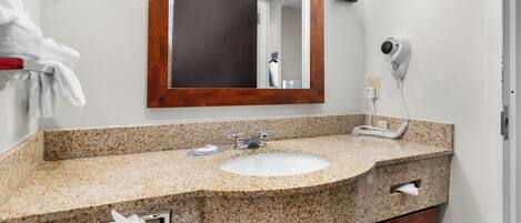 A bathroom with a granite countertop sink, a wooden-framed mirror, a hairdryer, and a towel rack.