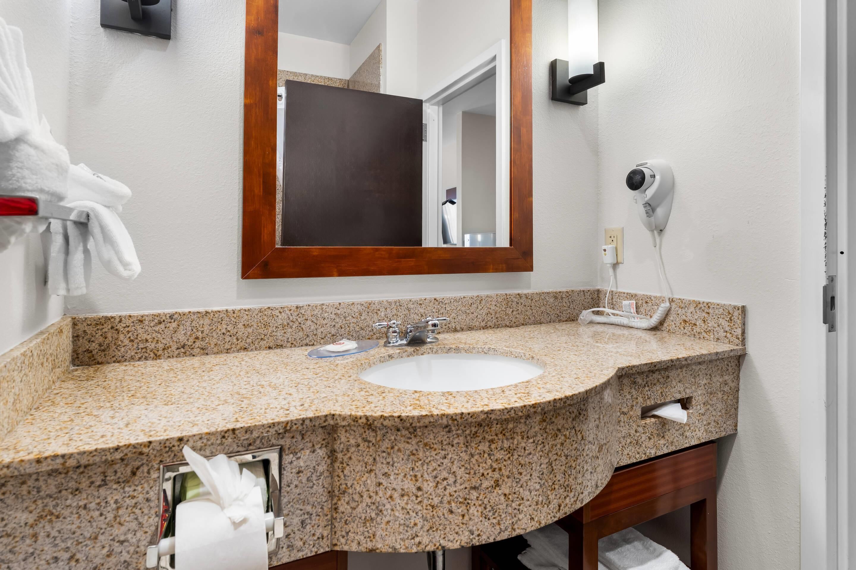 Baño con lavamanos de encimera de granito, espejo, toallero, secador de pelo y dispensador de toallas.