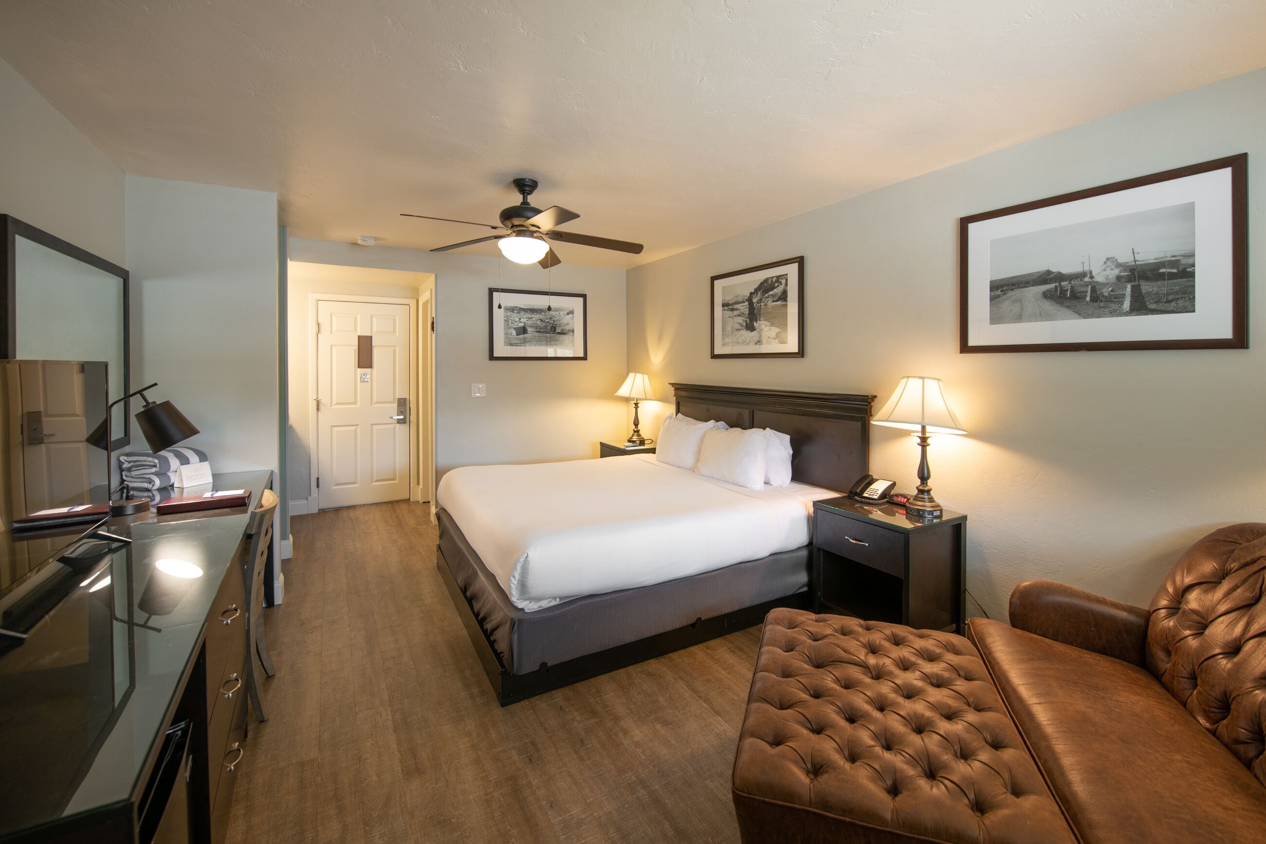 Une chambre d’hôtel avec un lit, un bureau, une chaise, un ventilateur de plafond et des tableaux encadrés accrochés au mur.